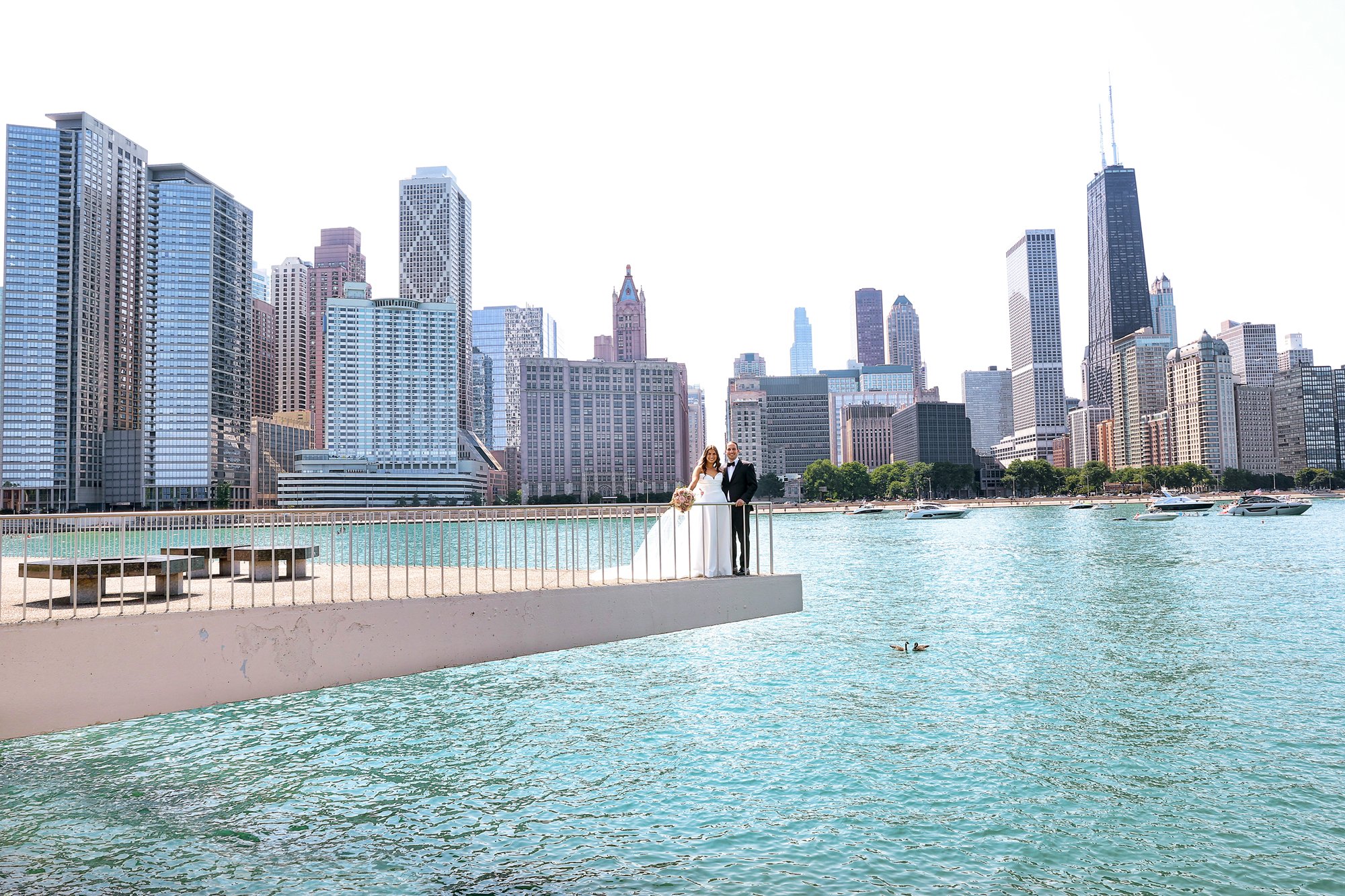 A bride and groom standing on a platform by the water with a city skyline in the background, including tall skyscrapers and boats on the water.