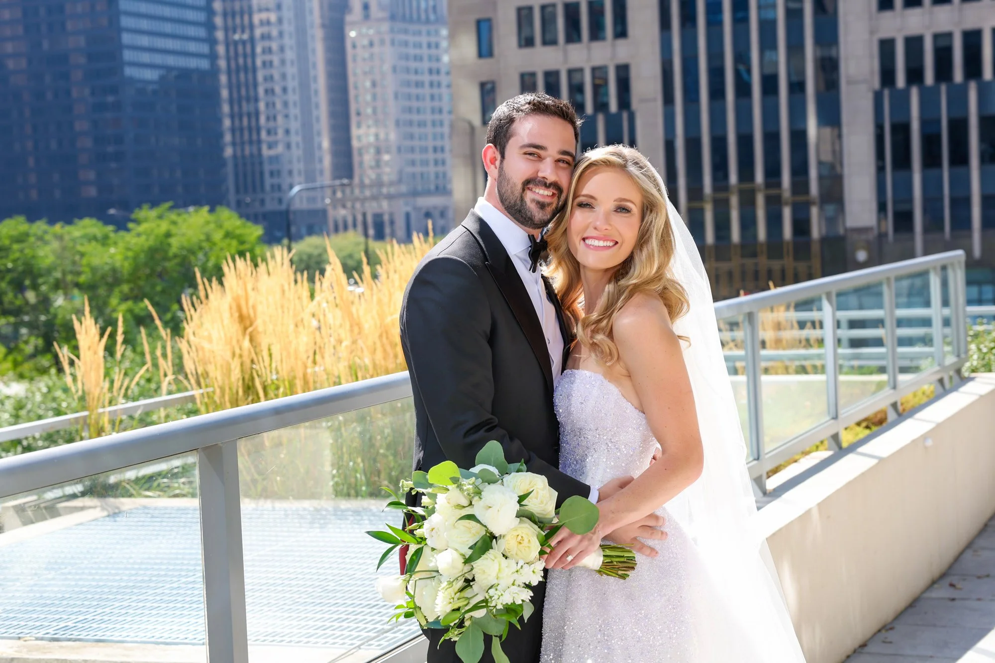A newlywed couple standing on a bridge outdoors in a city, smiling and holding a bouquet of white flowers, with tall buildings and greenery in the background.