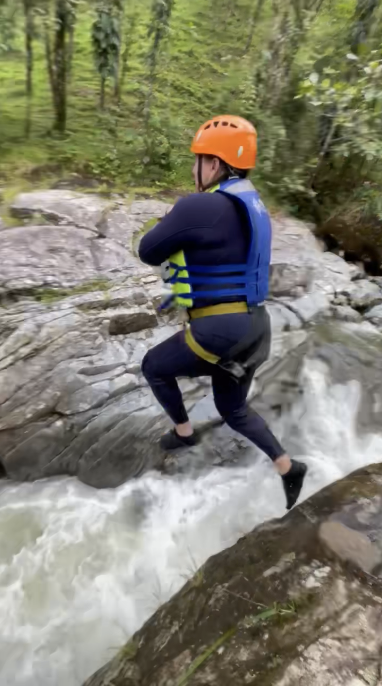 A person wearing an orange helmet, black wetsuit, and blue life vest crossing a rocky stream.