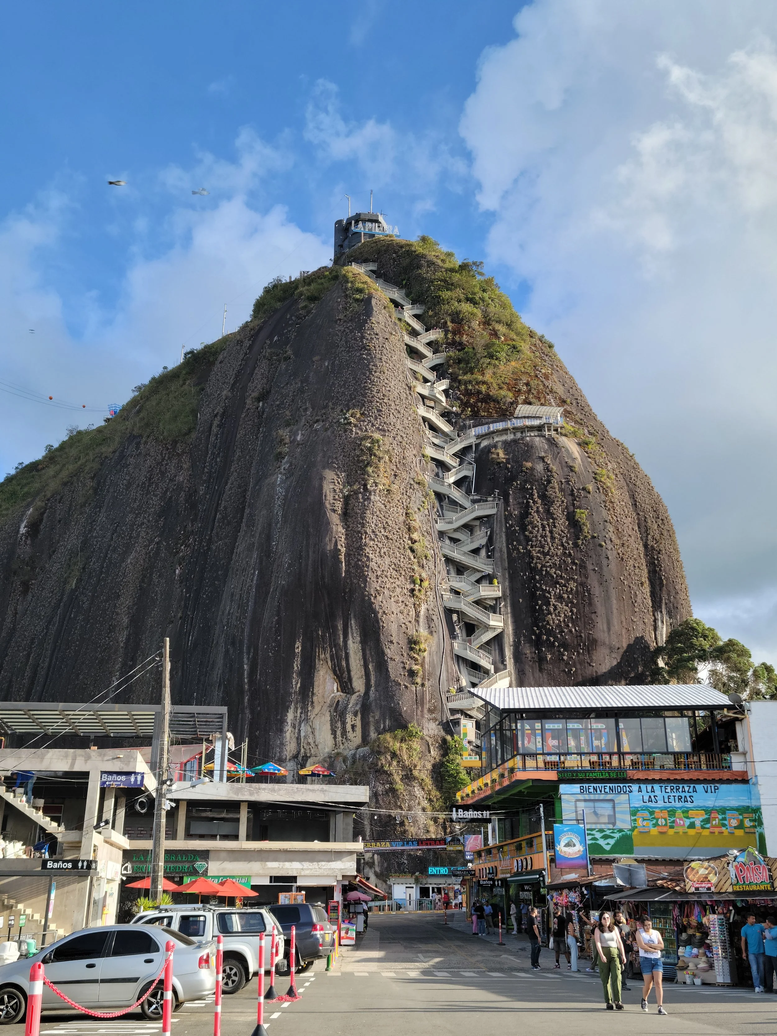 View of a large rocky hill with a staircase and a building at the top, with a busy street and shops at the base, and a bright blue sky with some clouds.