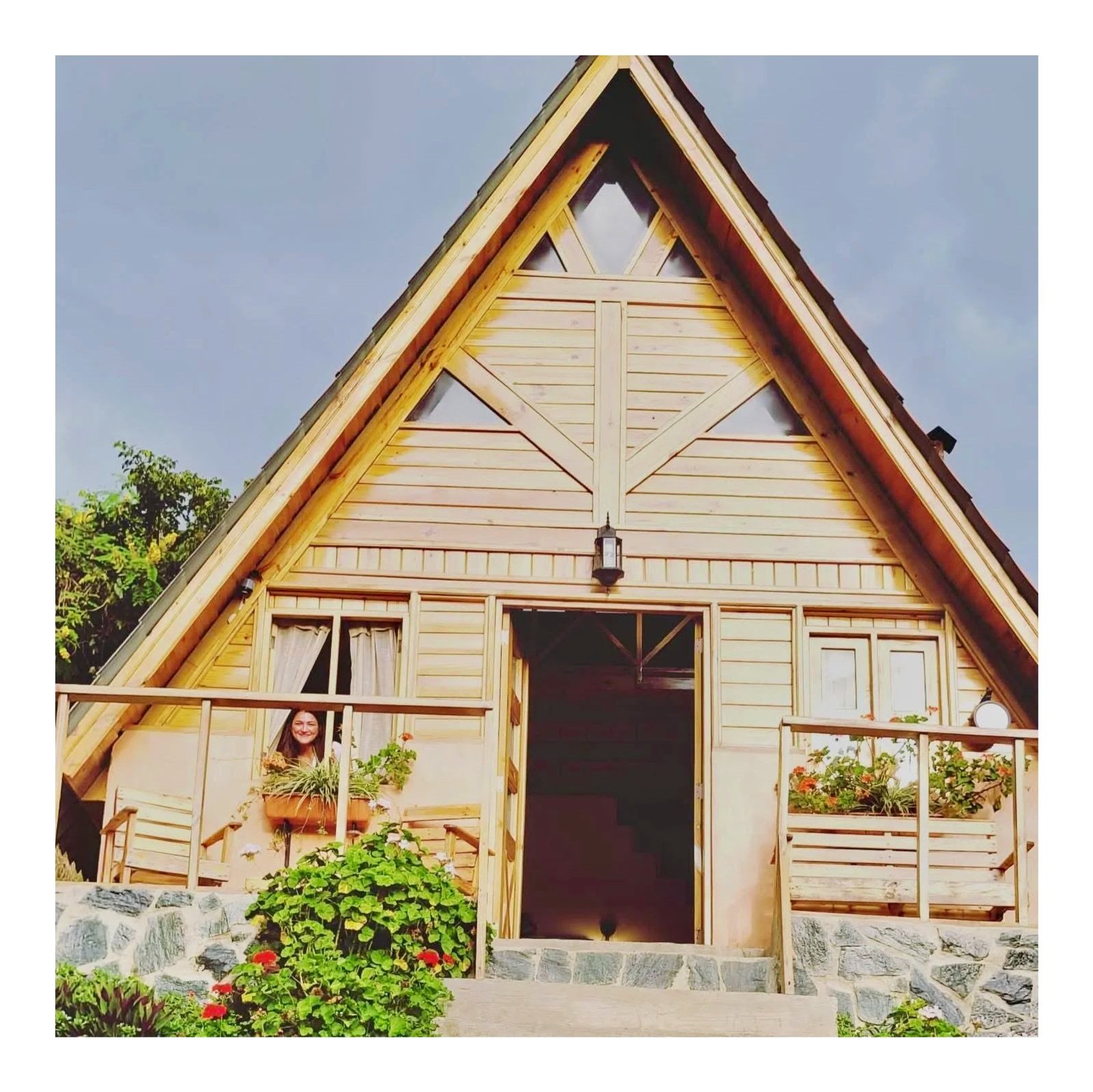 A wooden A-frame cabin with a small balcony and garden plants, a woman visible through a window, under a partly cloudy sky.