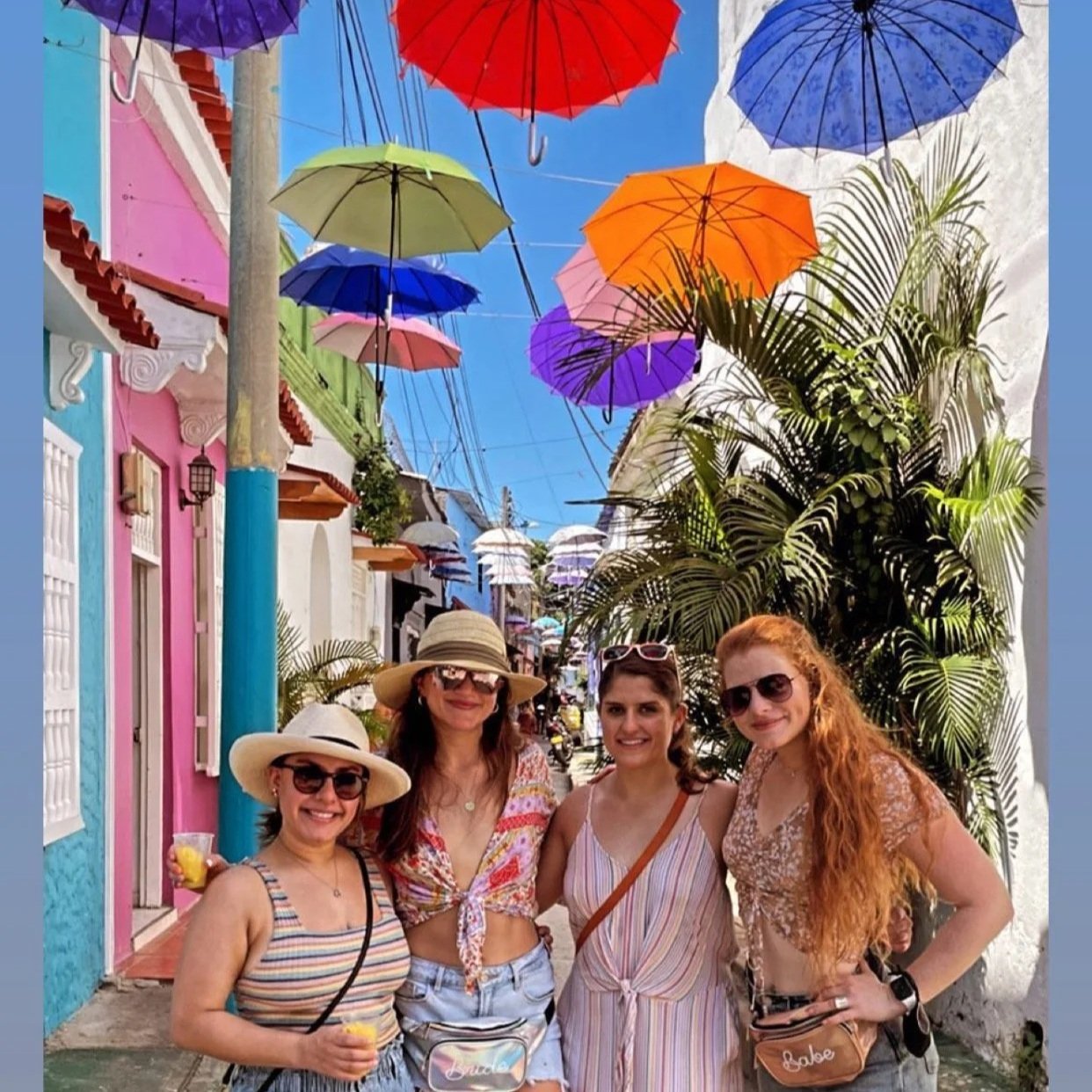 Four women smiling in a colorful street with hanging umbrellas and vibrant buildings.