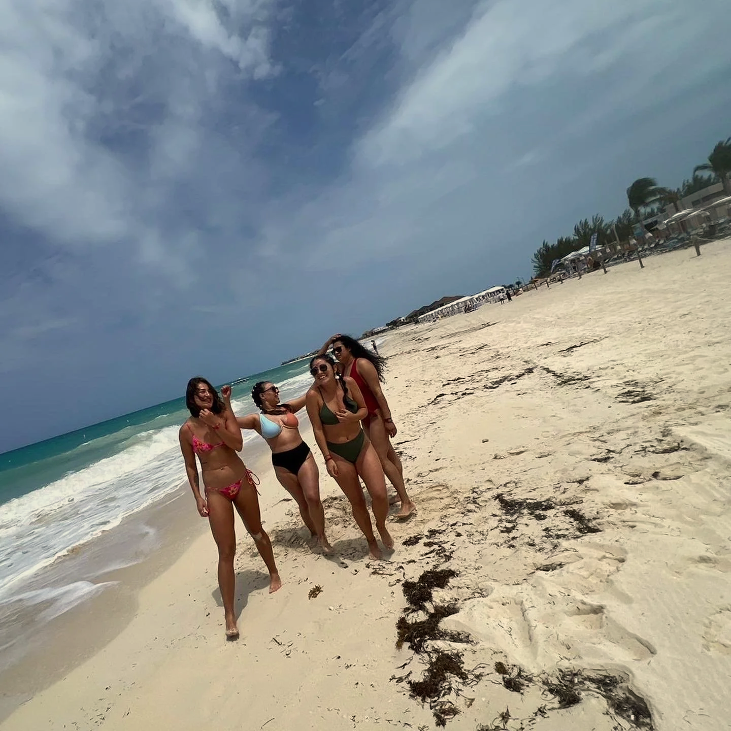 Group of four women in swimsuits walking along a sandy beach with palm trees and beach umbrellas in the background on a partly cloudy day.
