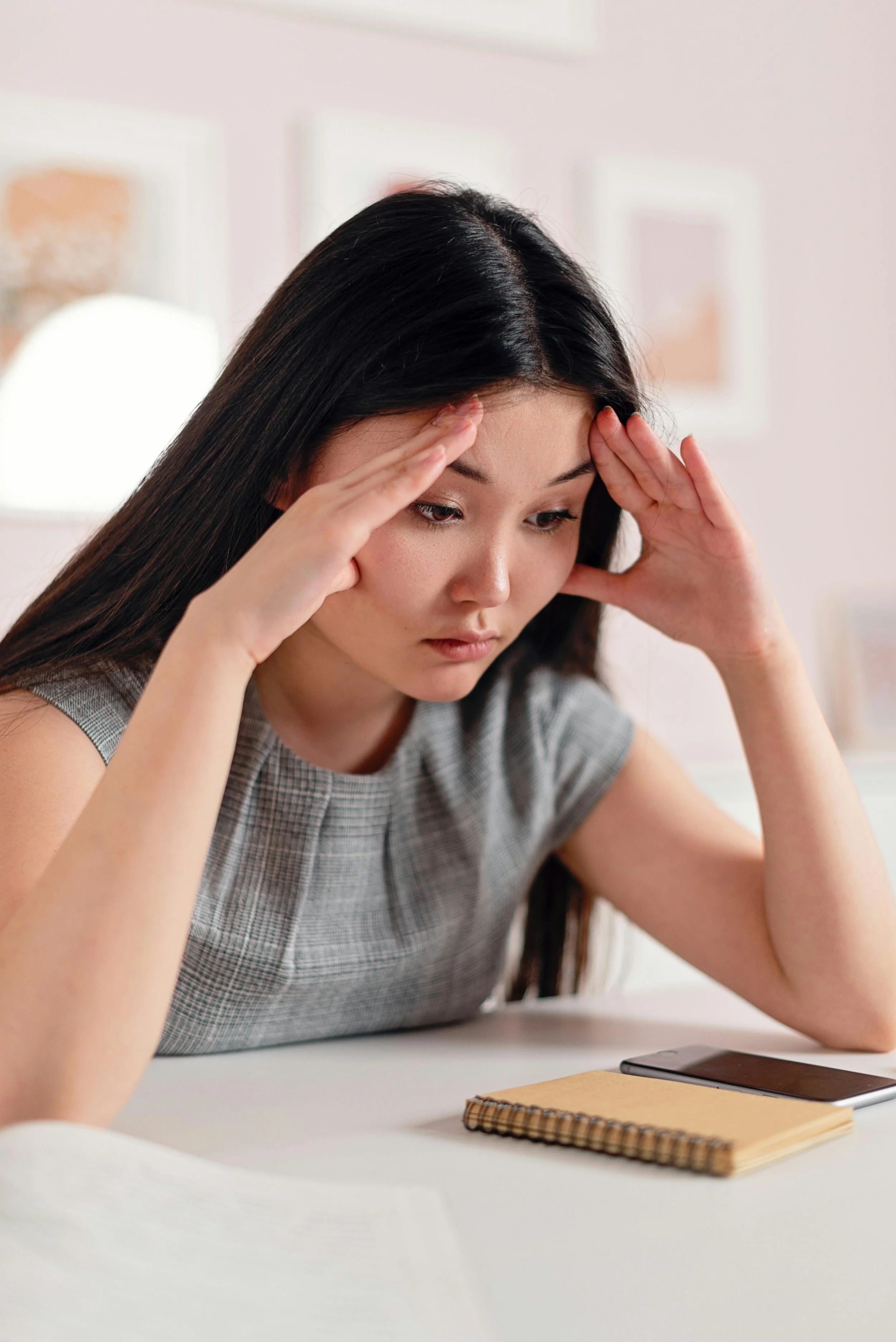 Young woman with long dark hair sitting at a desk with her hands on her forehead, appearing stressed or worried, with a smartphone, notebook, and paper in front of her.