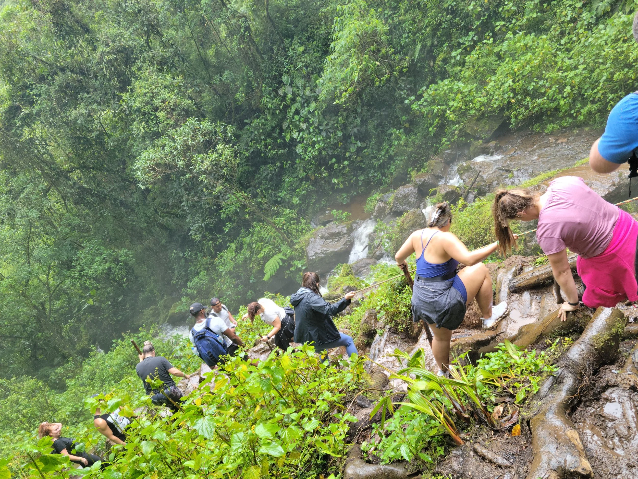 Group of people climbing over rocks and mud along a waterfall in a lush green forest.