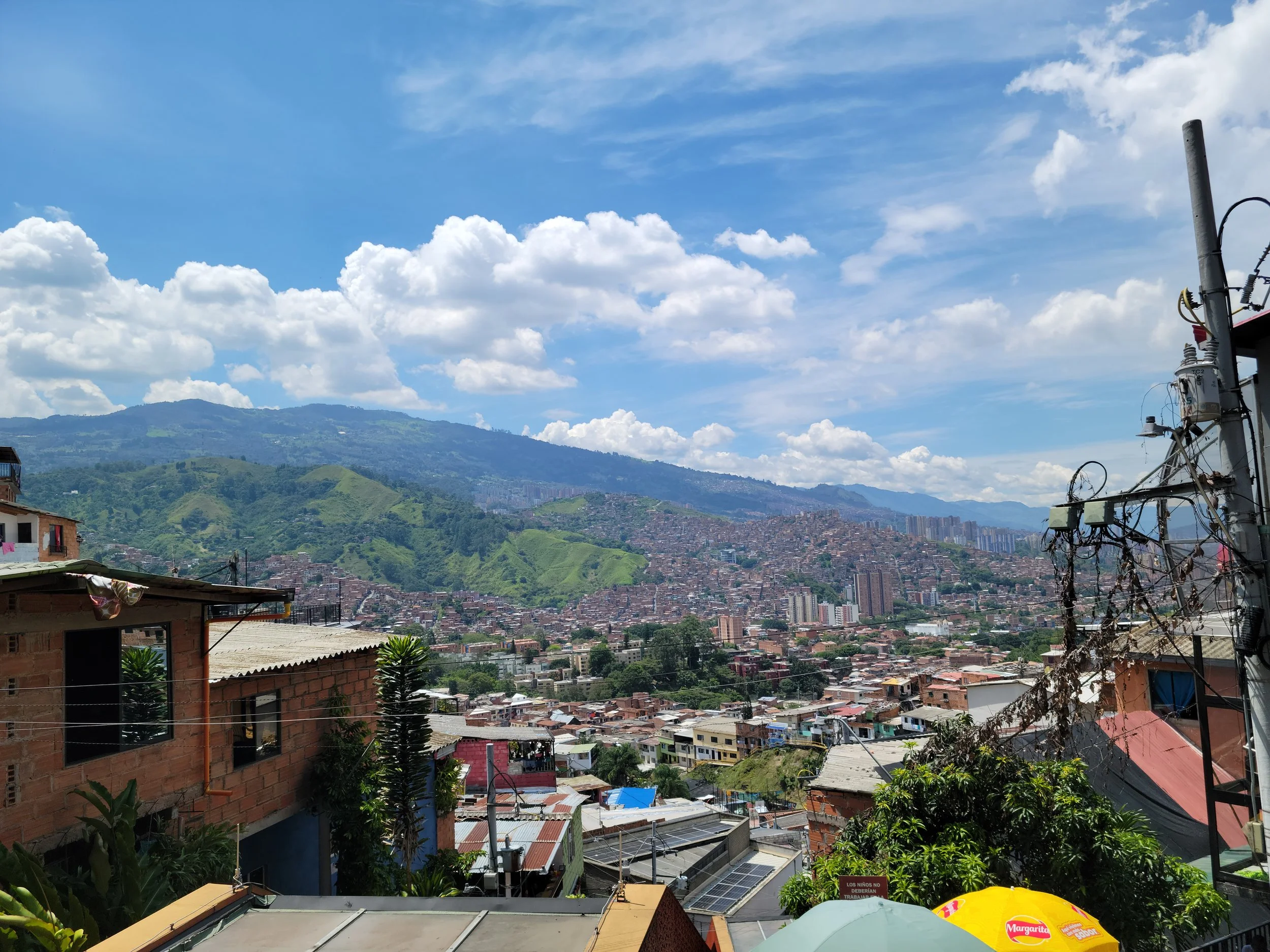 View of a city with densely packed buildings, green hills, and mountains in the background under a partly cloudy sky.