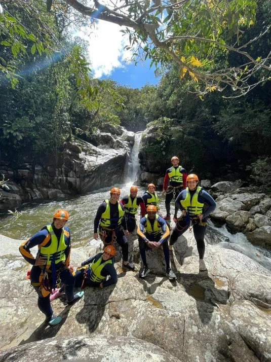 Group of eight people wearing life jackets and helmets standing on rocks beside a river with a small waterfall in the background, surrounded by greenery and trees under a partly cloudy sky.