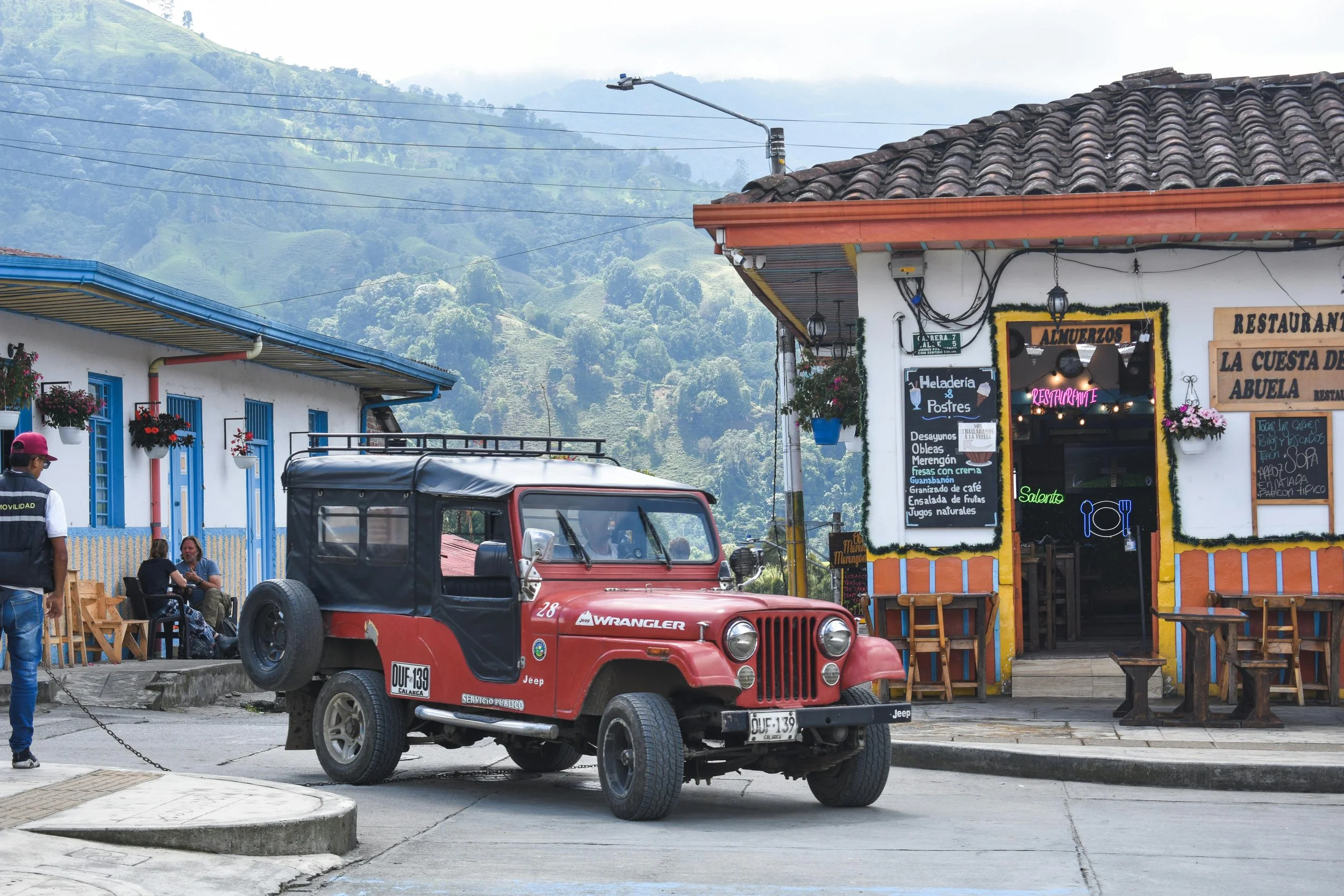 A red Jeep Wrangler parked in front of a restaurant in a mountainous area, with a person in a suit standing nearby and two people sitting outside the restaurant. The restaurant has colorful signage and hanging flowers.