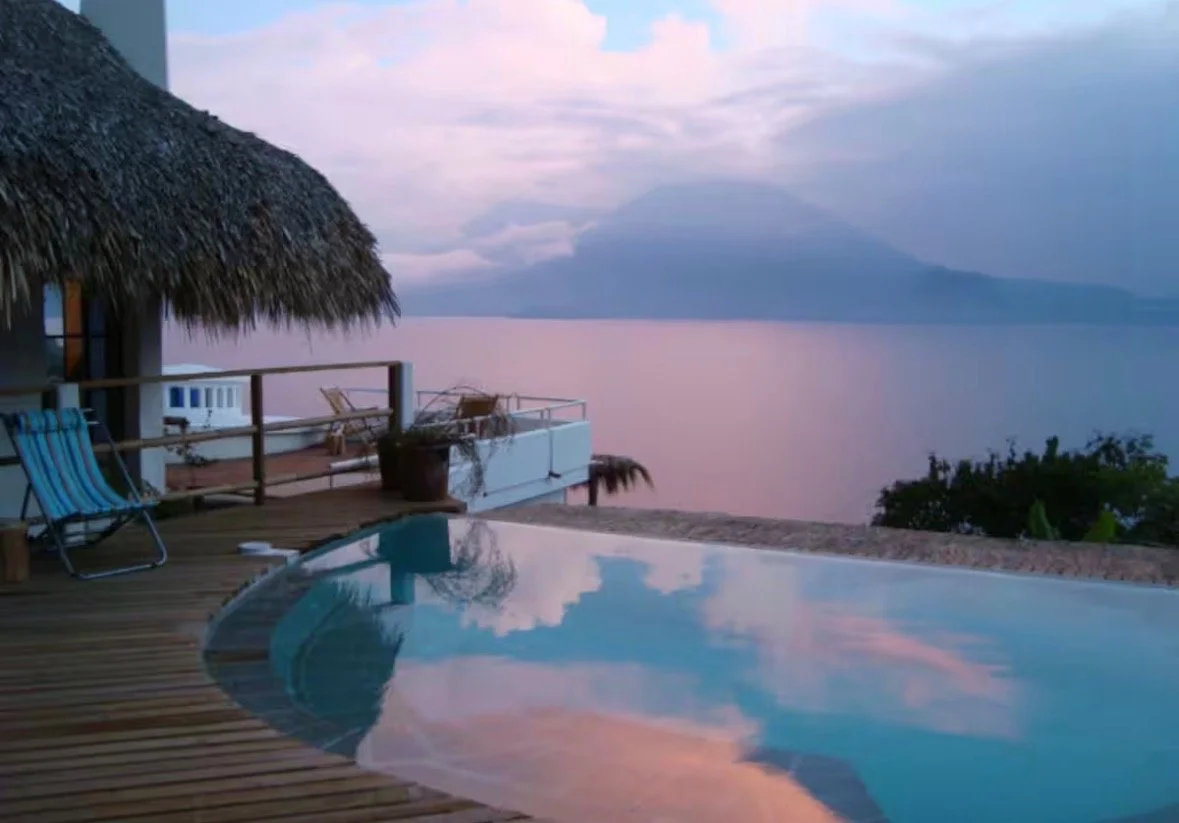 A poolside scene at sunset with a thatched roof hut, wooden deck chairs, potted plants, and a view of a calm body of water with mountains in the distance.
