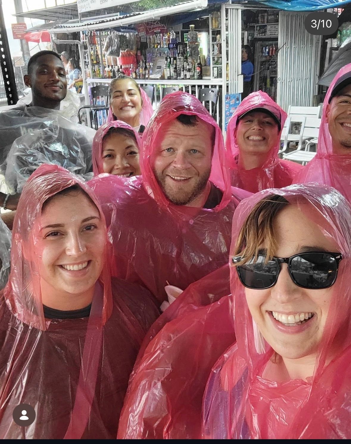 A group of smiling people wearing pink rain ponchos, some with sunglasses, in front of an outdoor bar or café during rainy weather.