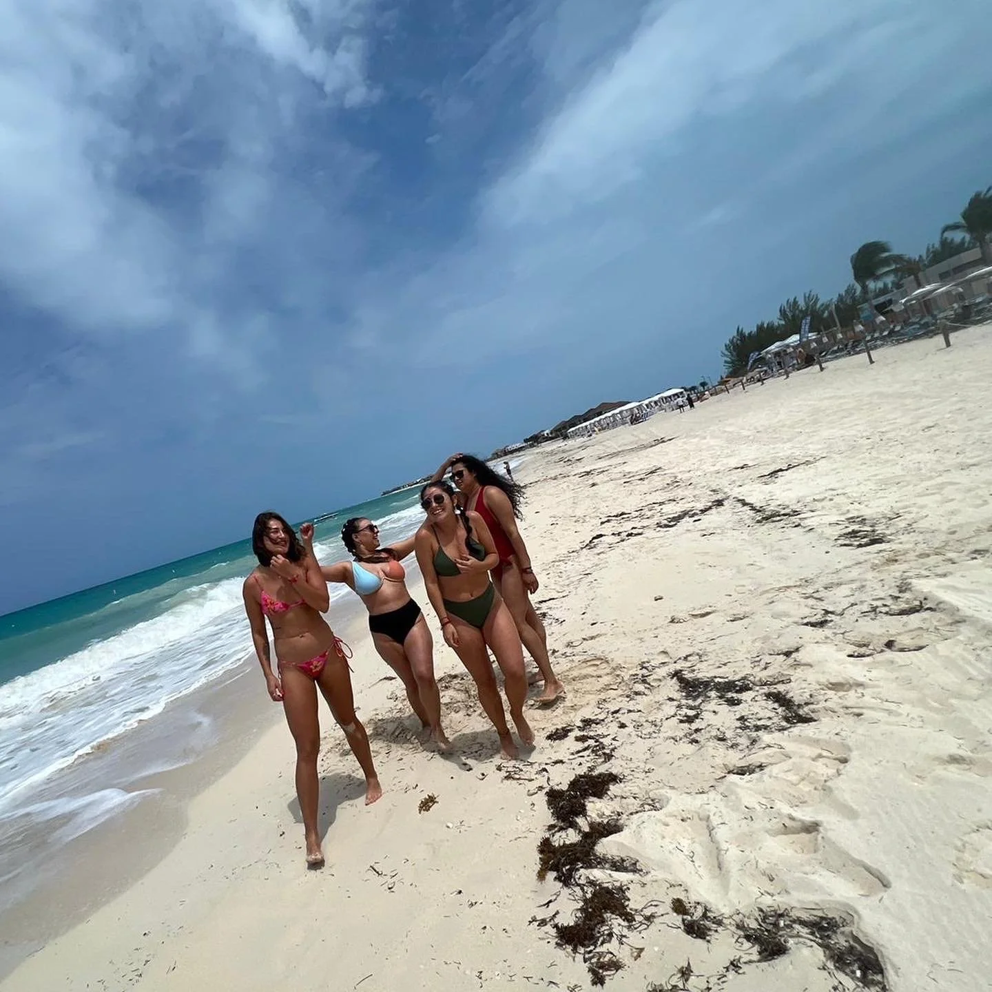 Four women in swimsuits standing together on a sandy beach with ocean waves and a blue sky in the background.
