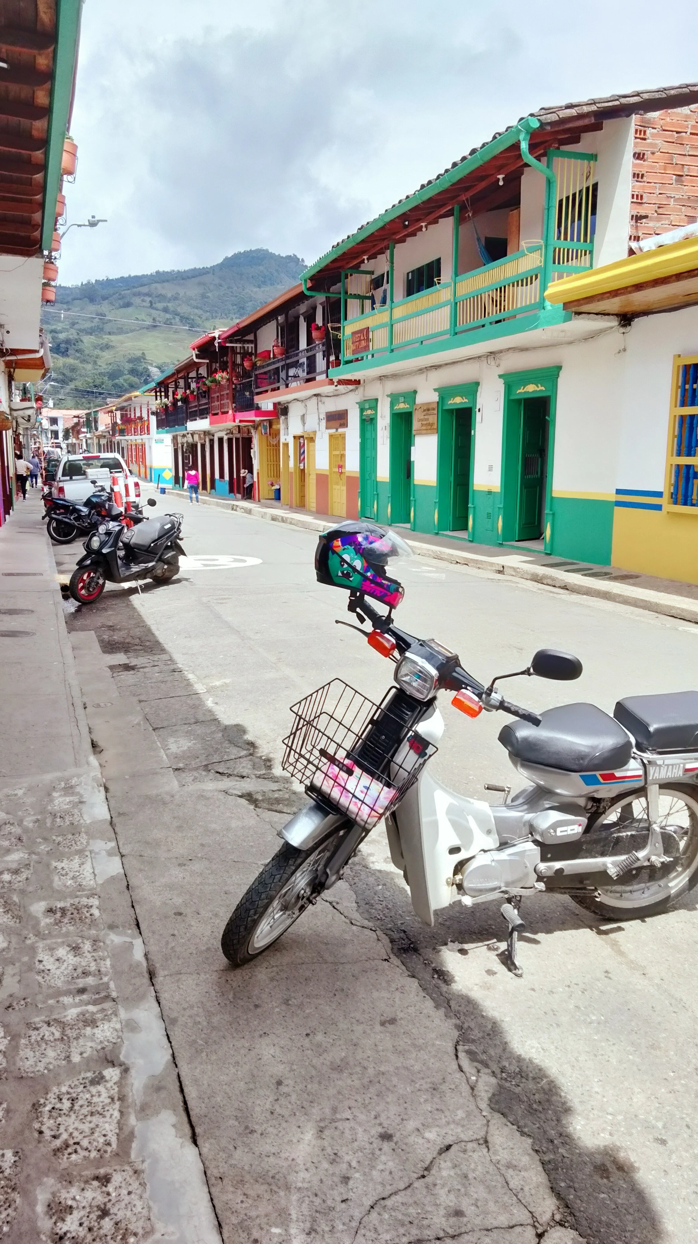 Colorful buildings with balconies lining a street, parked motorcycles, and a scooter with a helmet on handlebar in a town with mountainous background.
