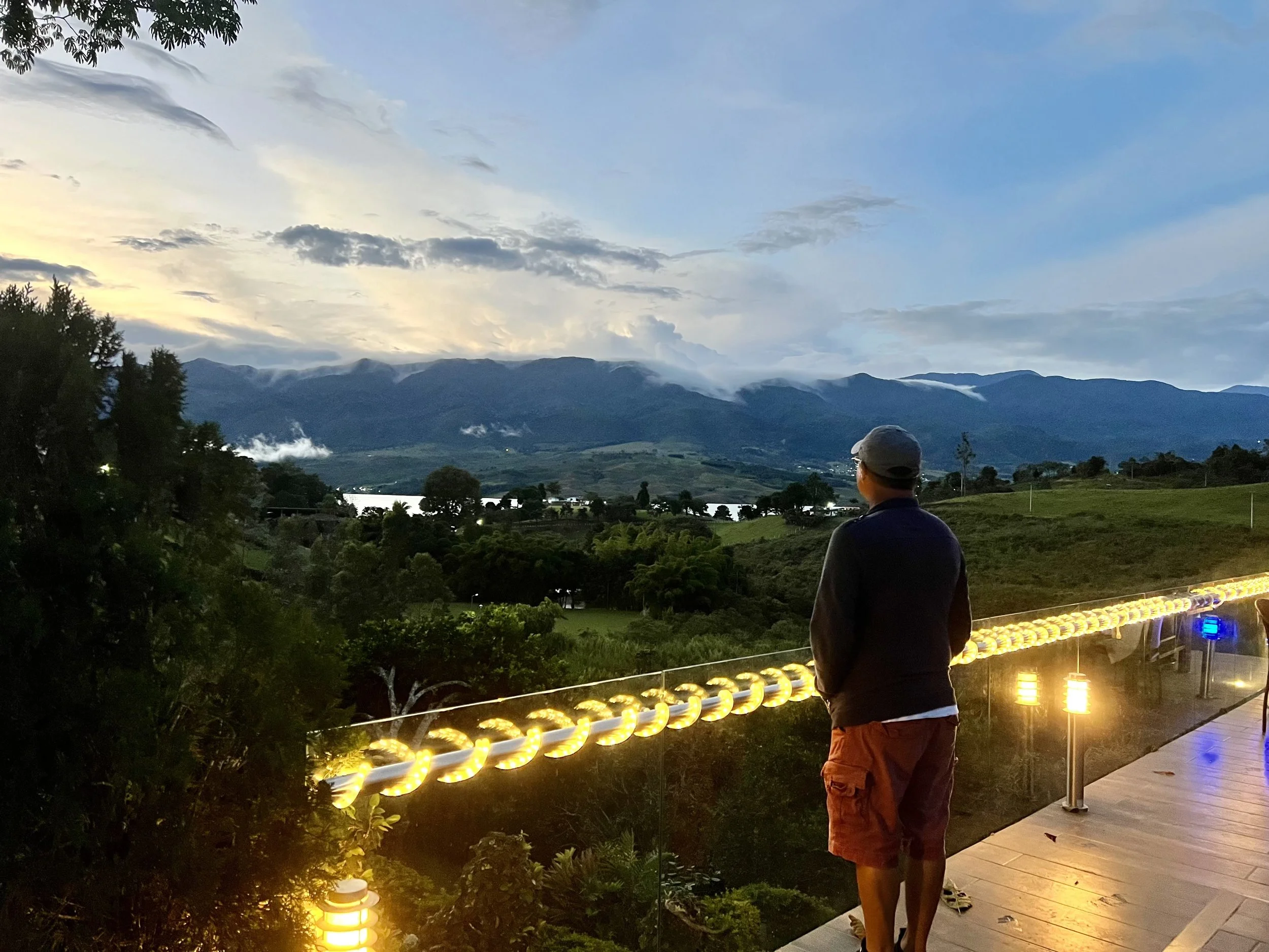A man standing on a terrace overlooking a scenic landscape with mountains, a lake, and a partly cloudy sky at dusk. The terrace has yellow and blue lights.