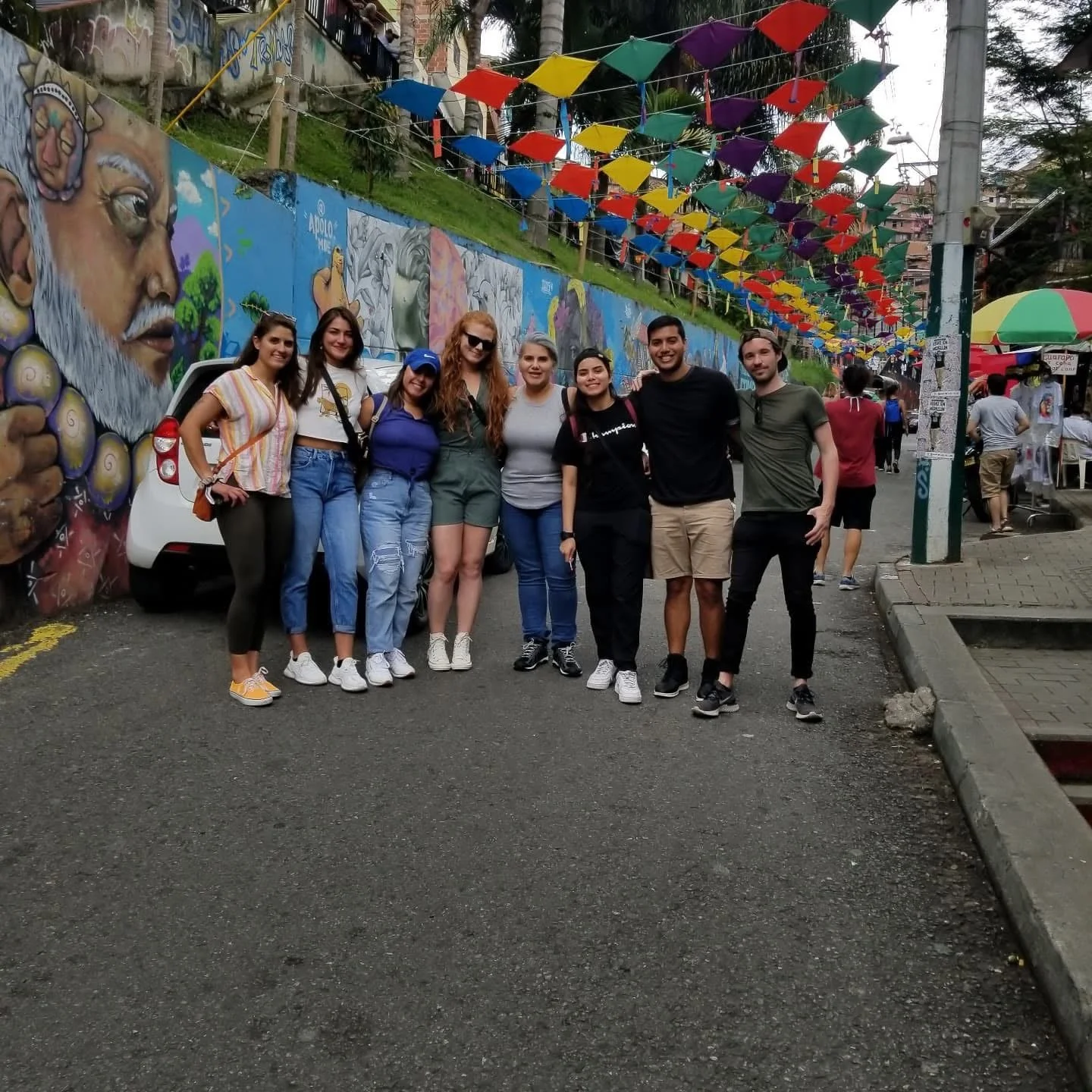 A group of nine people standing together on a street decorated with colorful umbrellas and murals, smiling for the photo.