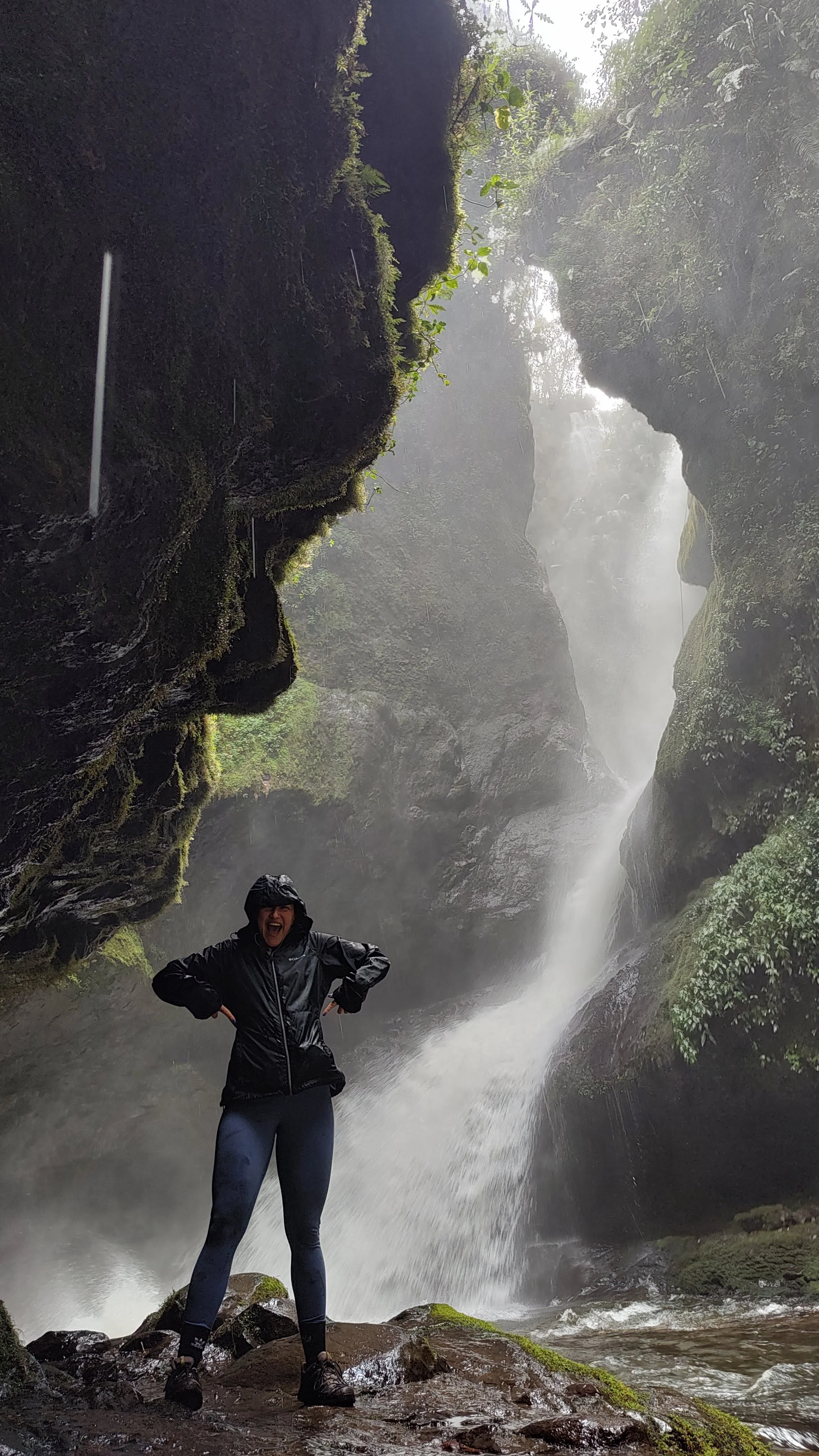A person in a black rain jacket and leggings standing on rocks inside a canyon with a waterfall in the background, surrounded by moss-covered rocks and lush greenery.