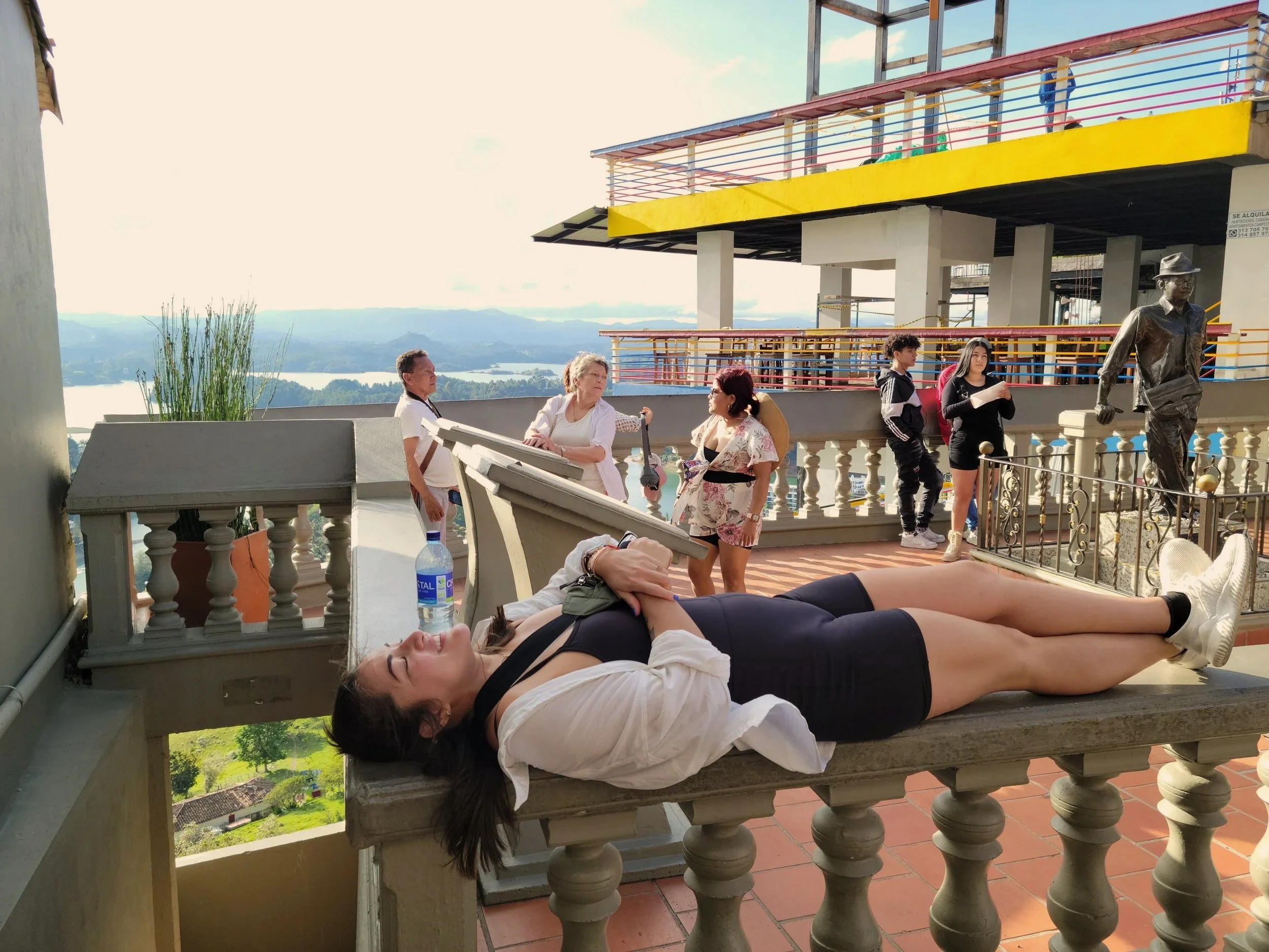 A woman lies on a railing with her eyes closed, holding a water bottle, while people stand and chat in the background on a rooftop with scenic views and a modern building.