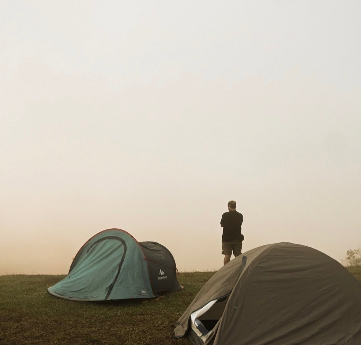 A person standing on a grassy hilltop with their arms crossed, facing away from the camera, surrounded by two tents, with a hazy sky in the background.