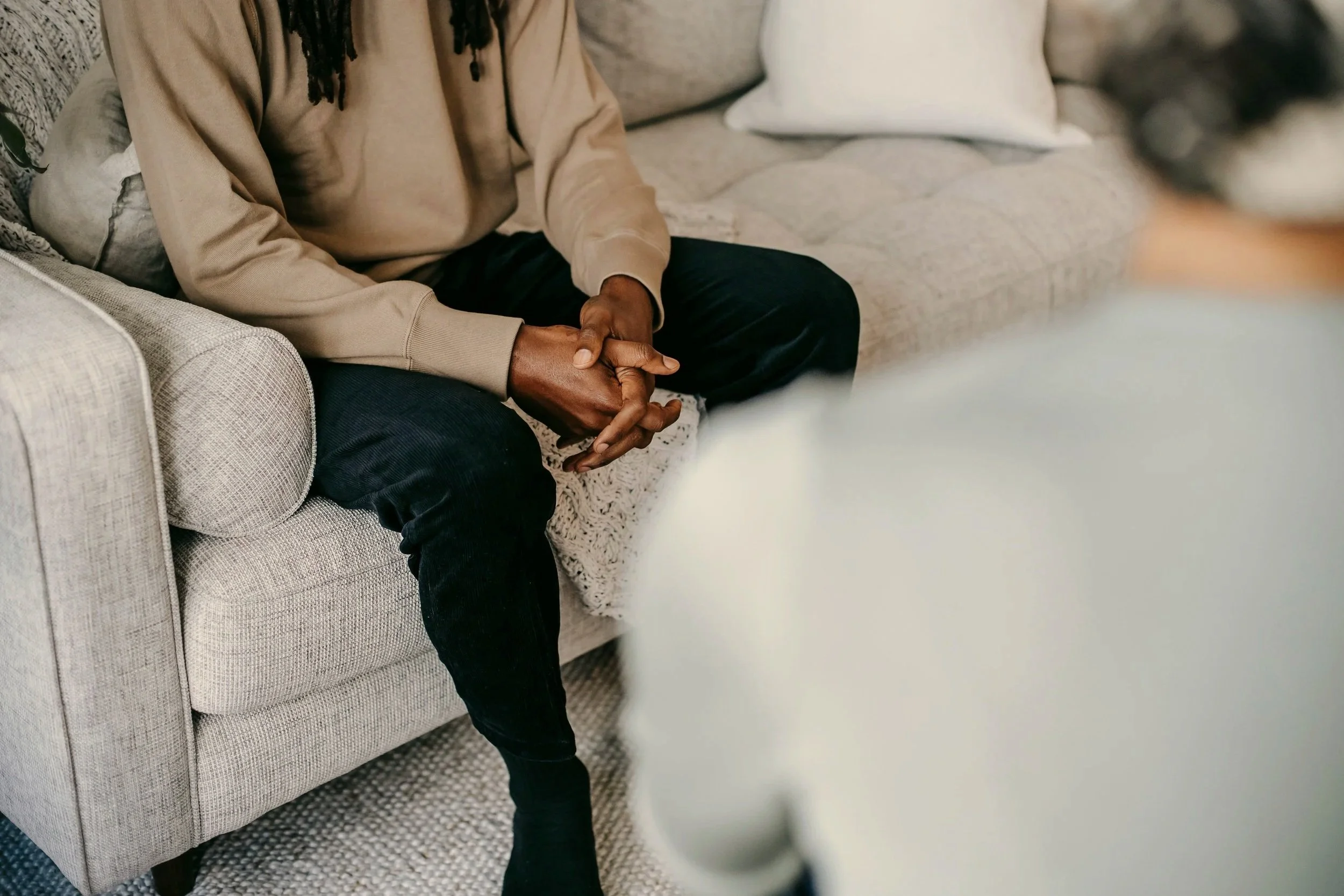 Person with dark skin wearing a beige hoodie and black pants sitting on a light-colored sofa with their hands clasped together.