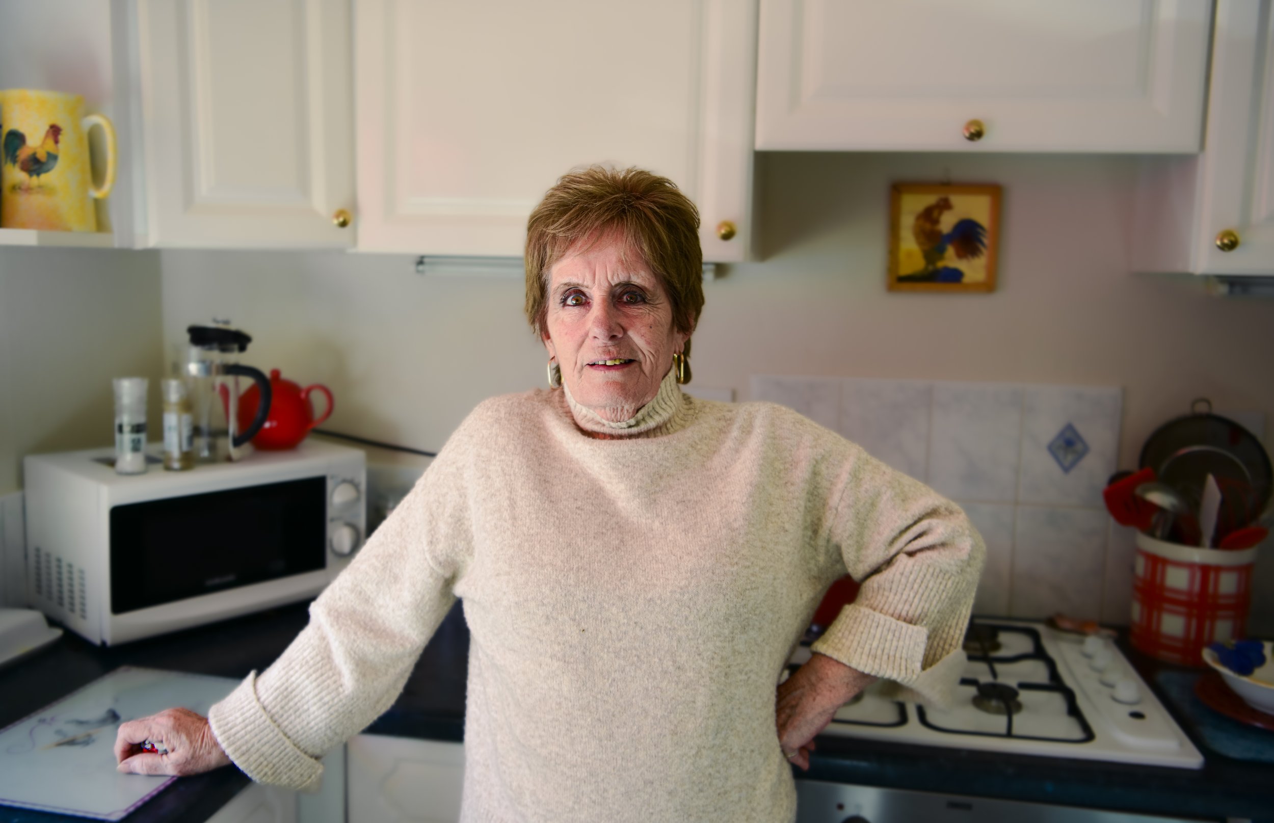 An older woman with short brown hair, wearing a beige sweater, standing in a kitchen with her hand on the countertop, making a surprised or confused expression.