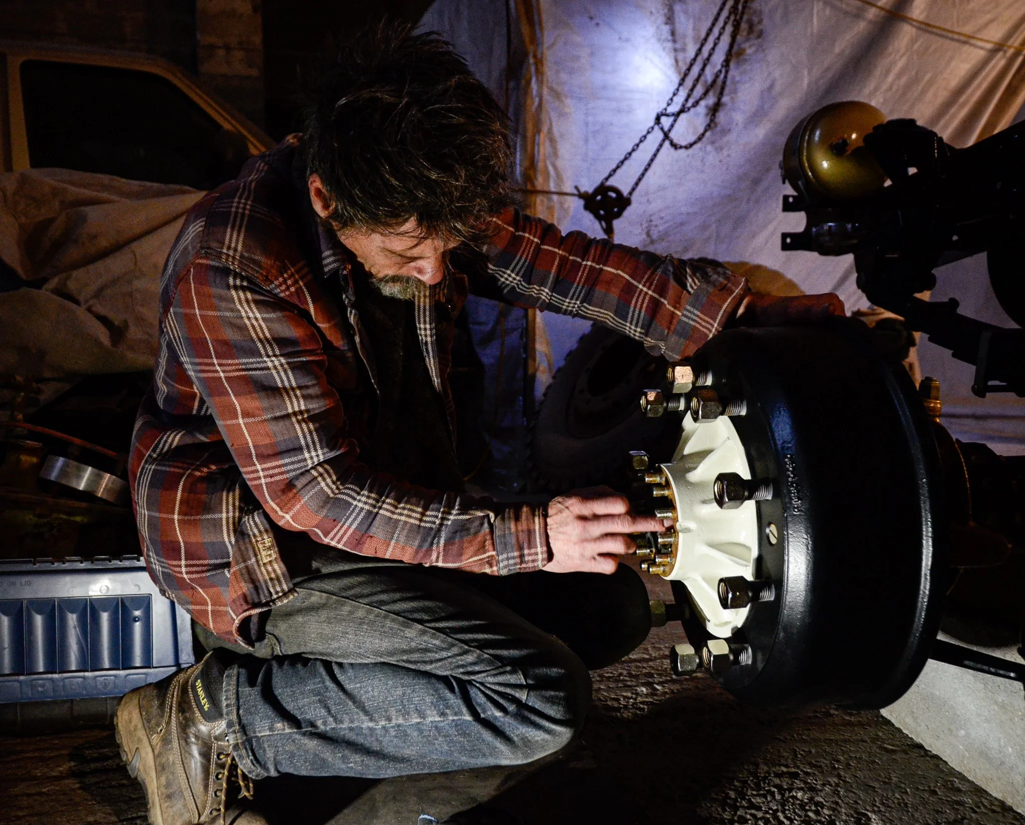 A man wearing a plaid shirt is working on the wheel of a vehicle, tightening or loosening bolts, in a garage or workshop.
