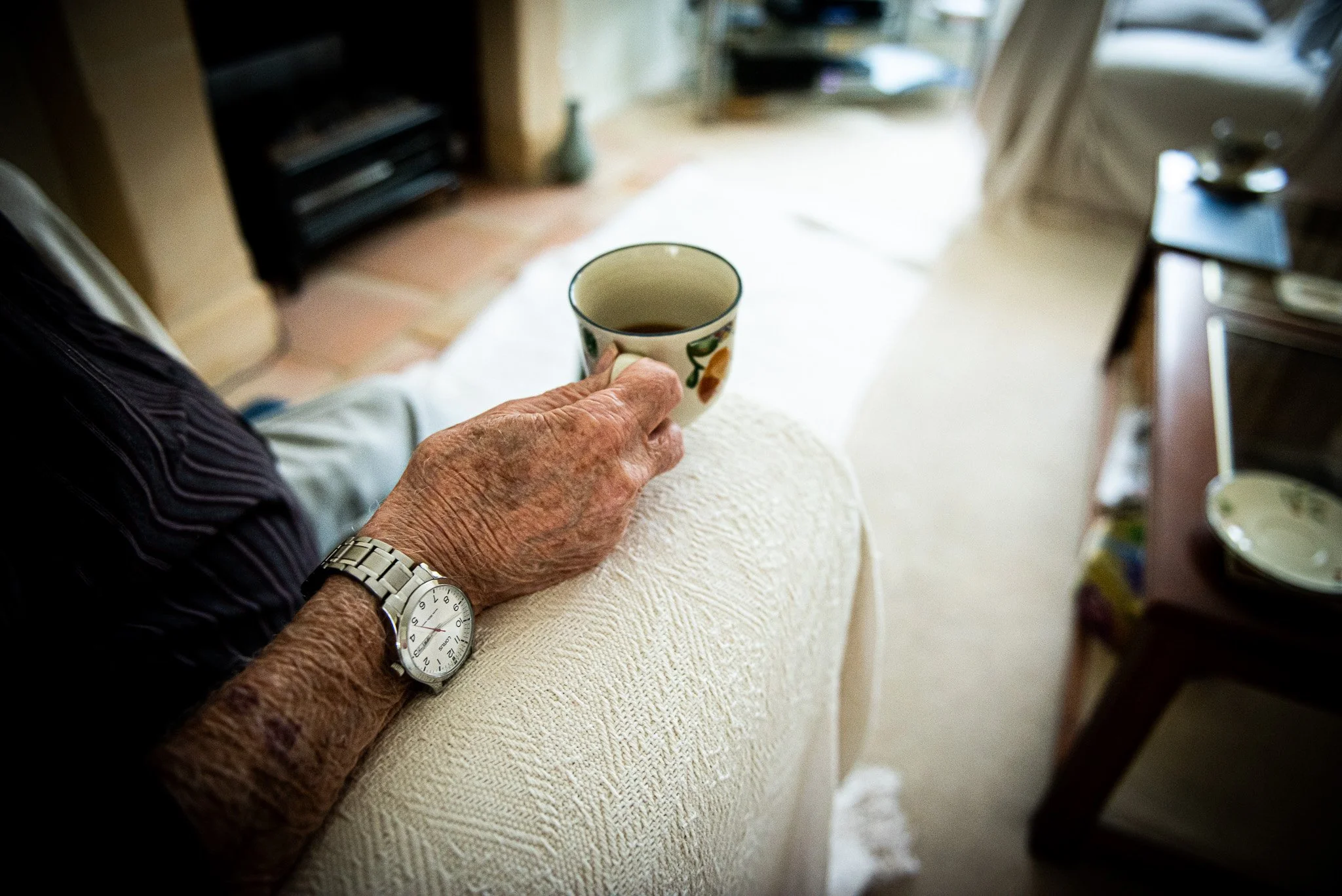 Close-up of an elderly person's hand holding a ceramic cup filled with a hot beverage, resting on their knee covered with a beige textured blanket, in a cozy living room.
