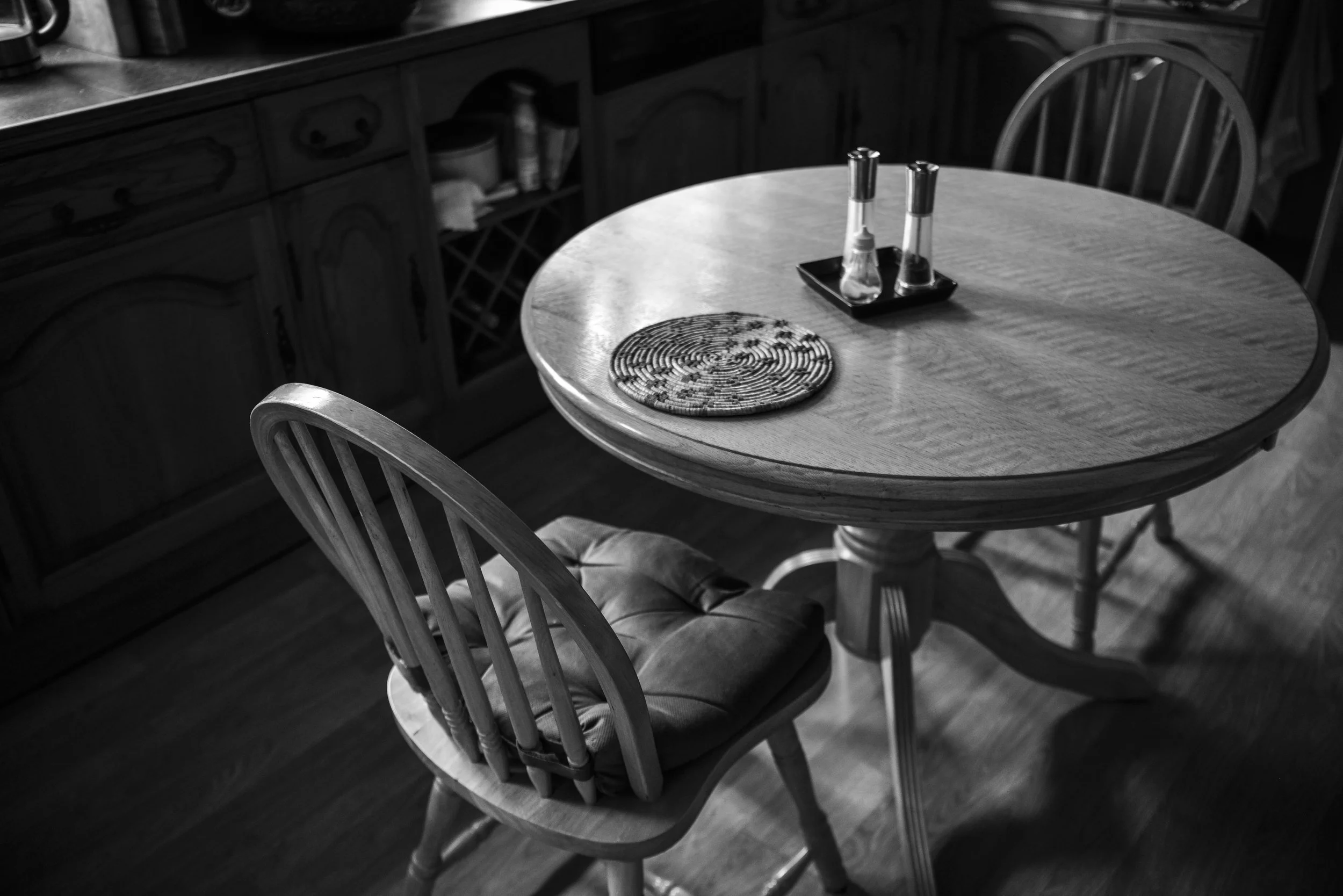 A round wooden dining table with a cushion on one of the chairs, a cloth placemat, and salt and pepper shakers in a small tray.