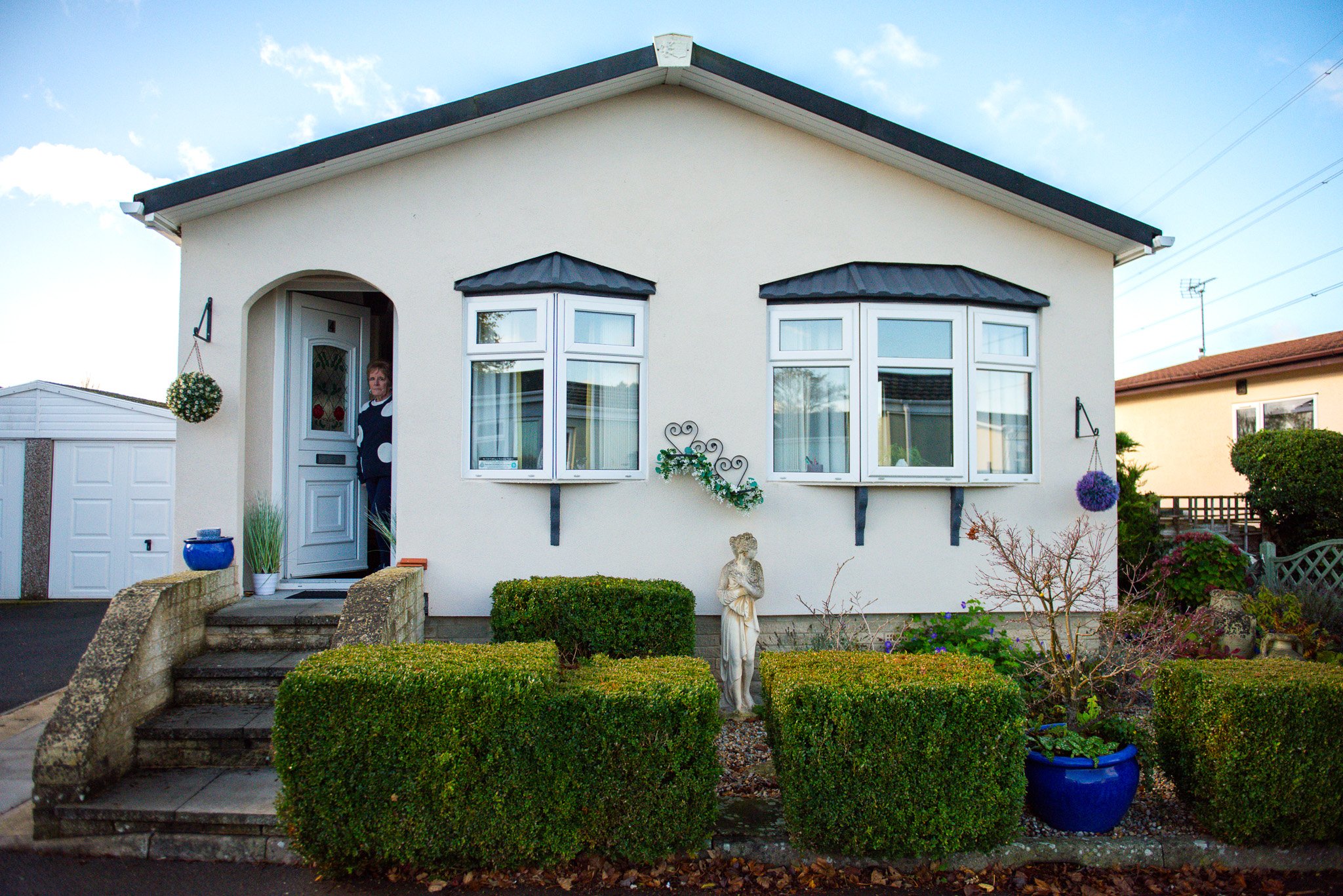 Front view of a white house with small garden and hedges, a statue of a woman, and a woman standing by the door.