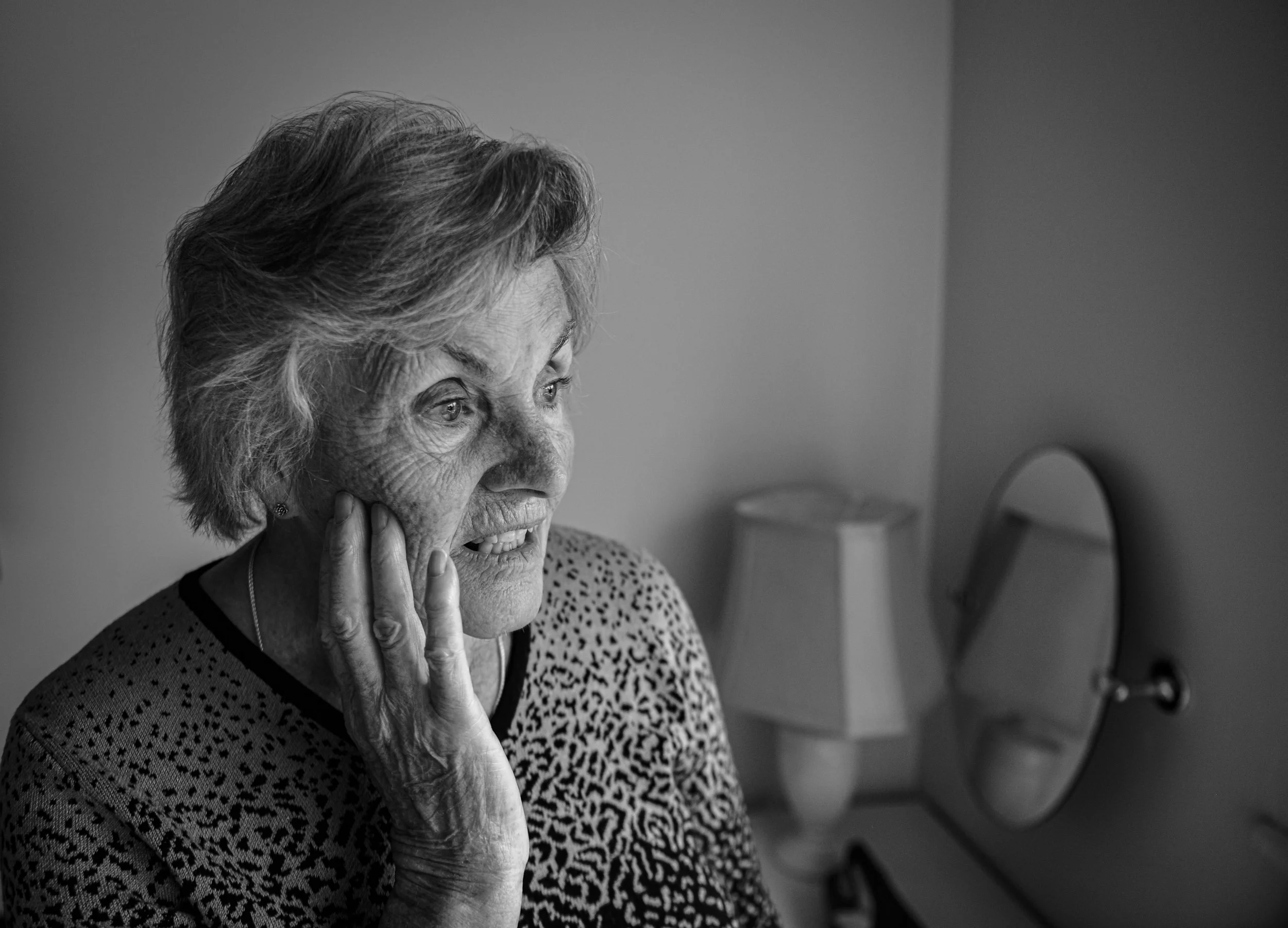 An elderly woman with short, wavy hair and a patterned top is looking into a mirror with a concerned or distressed expression, touching her face with her hand.