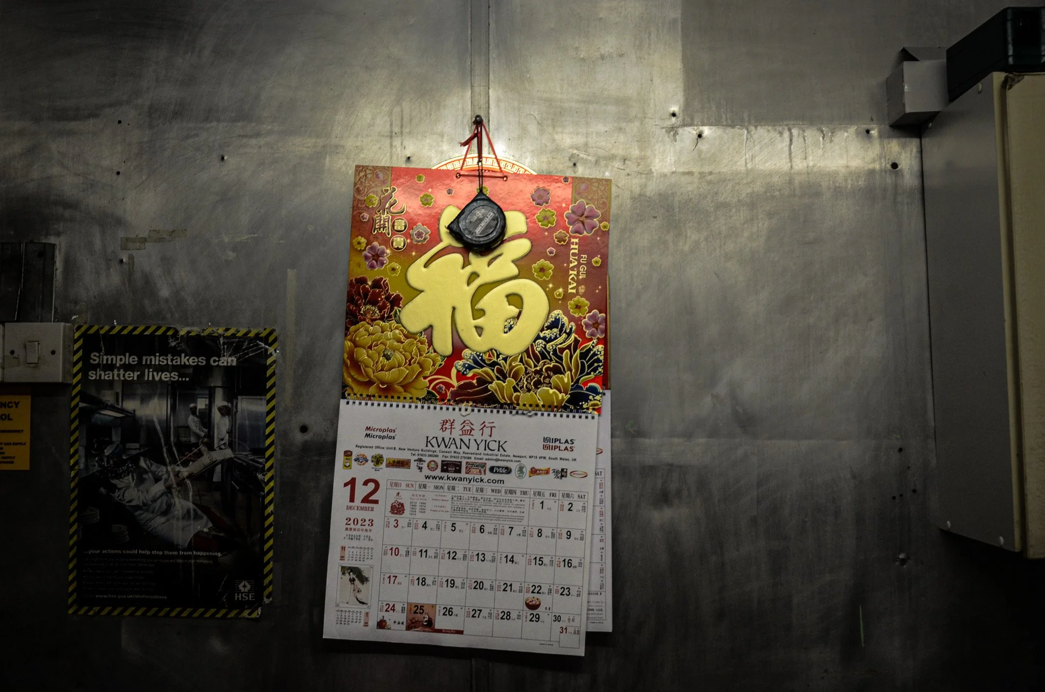 A wall calendar for December 2023 hanging on a metal wall with a decorative Chinese New Year poster above it and a black item hanging from the top of the poster. The calendar shows the days and dates, and the poster has a large Chinese character with
