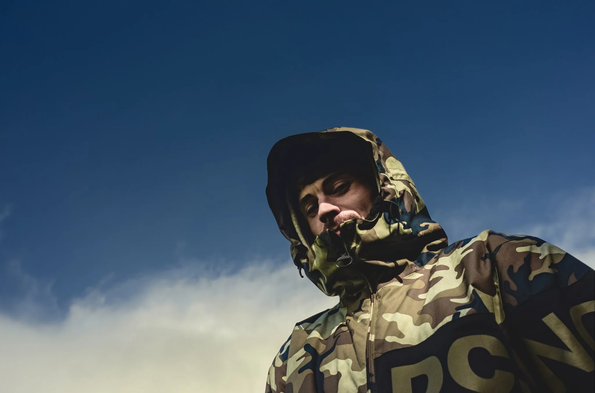 Person wearing camouflage jacket with hood, looking down, against a blue sky with some clouds.
