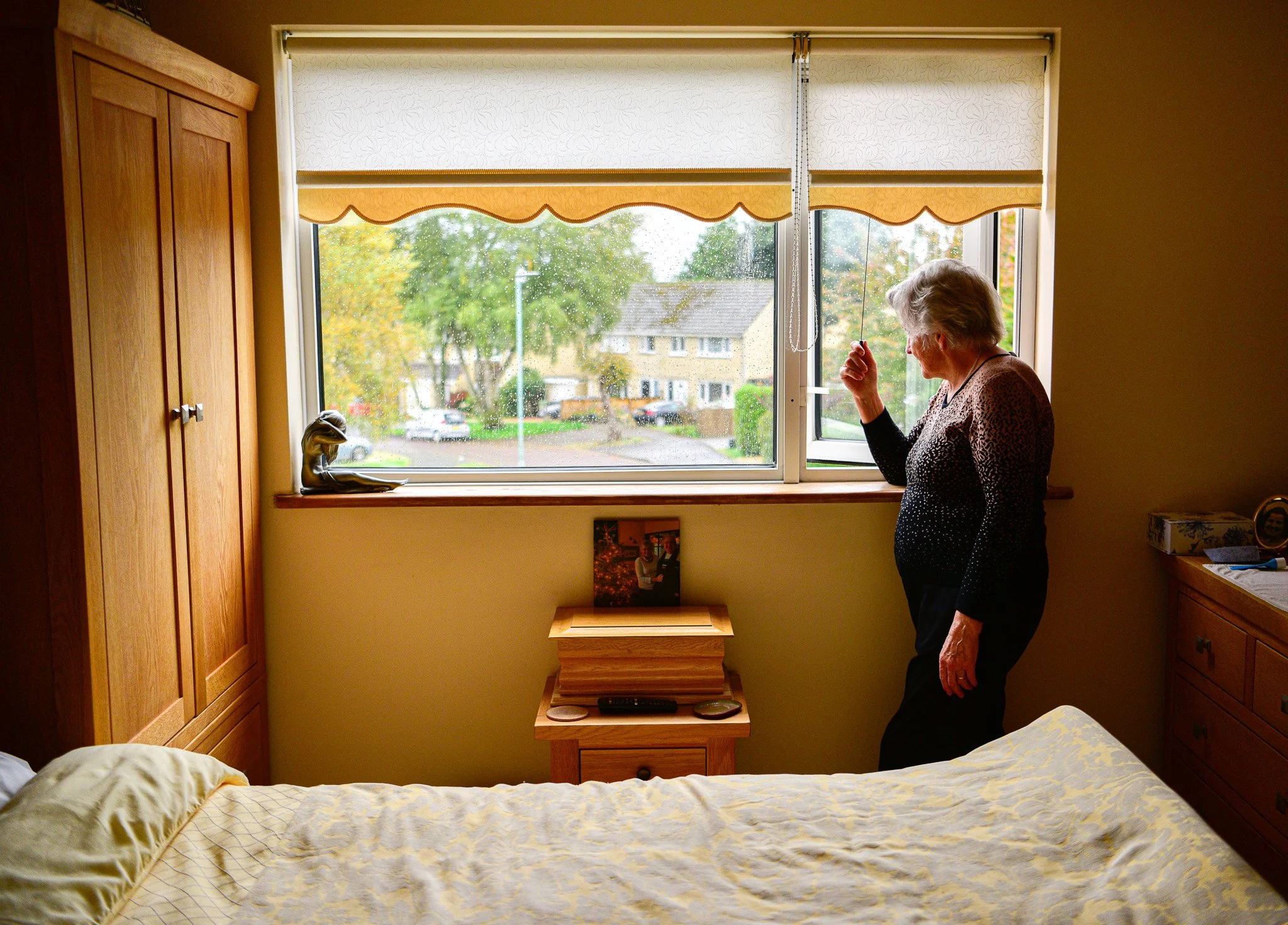 An elderly woman with short gray hair stands by an open window in a bedroom, looking outside on a rainy day. The room has wooden furniture, including a wardrobe, a bedside table, and a dresser. A small picture and a statue are on the windowsill, and 
