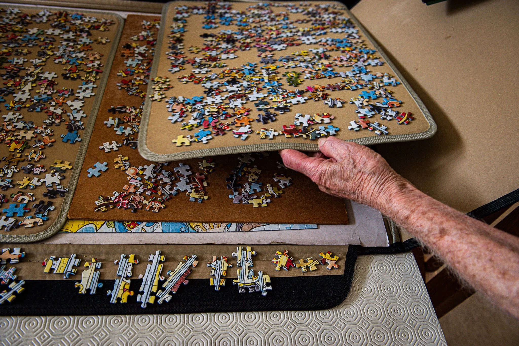 An elderly person's hand holds a board with scattered puzzle pieces, with more puzzle pieces spread on a table and puzzle boards in the background.