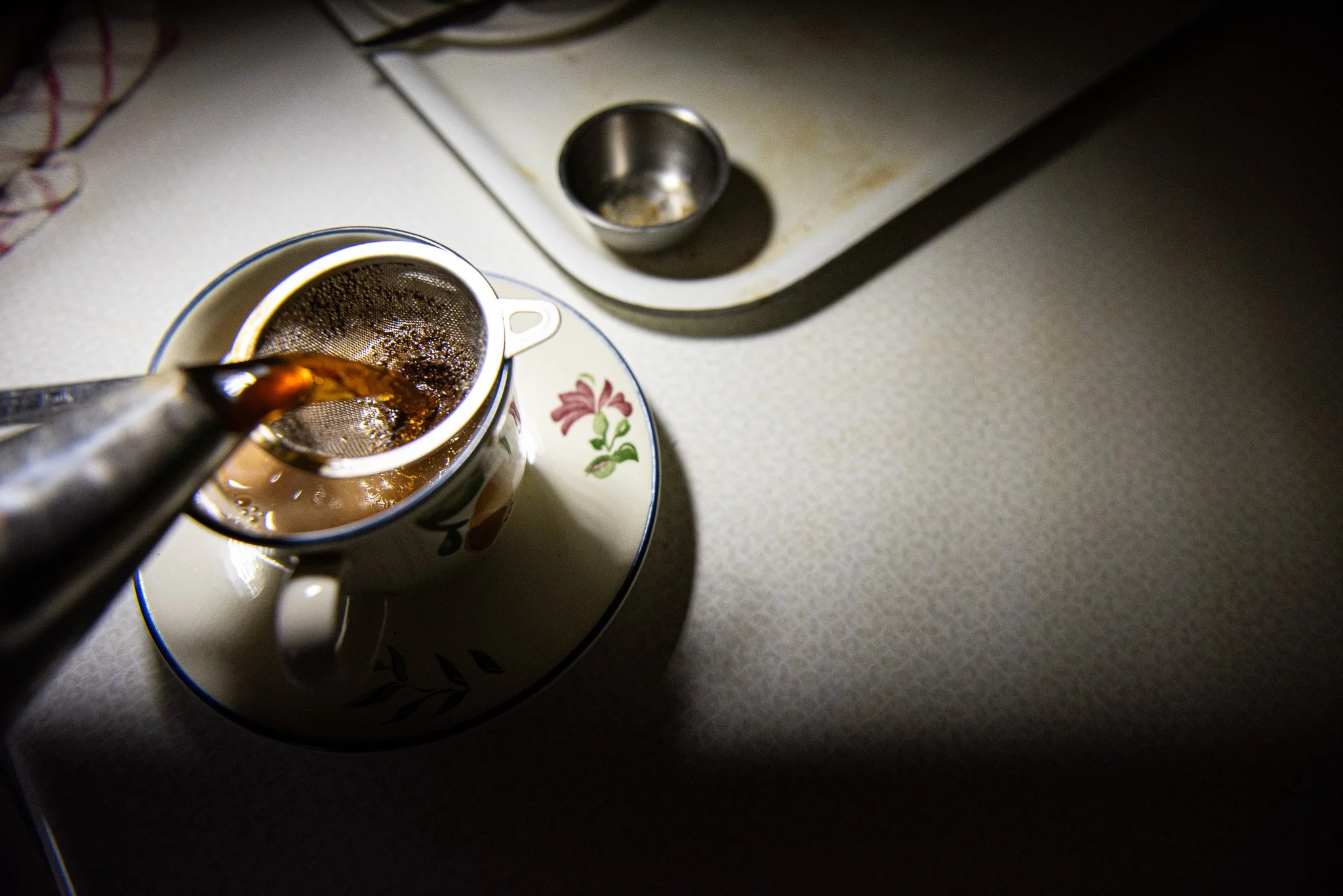 Pouring hot coffee through a metal infuser into a floral mug placed on a matching saucer, with a small empty metal cup on a tray nearby.