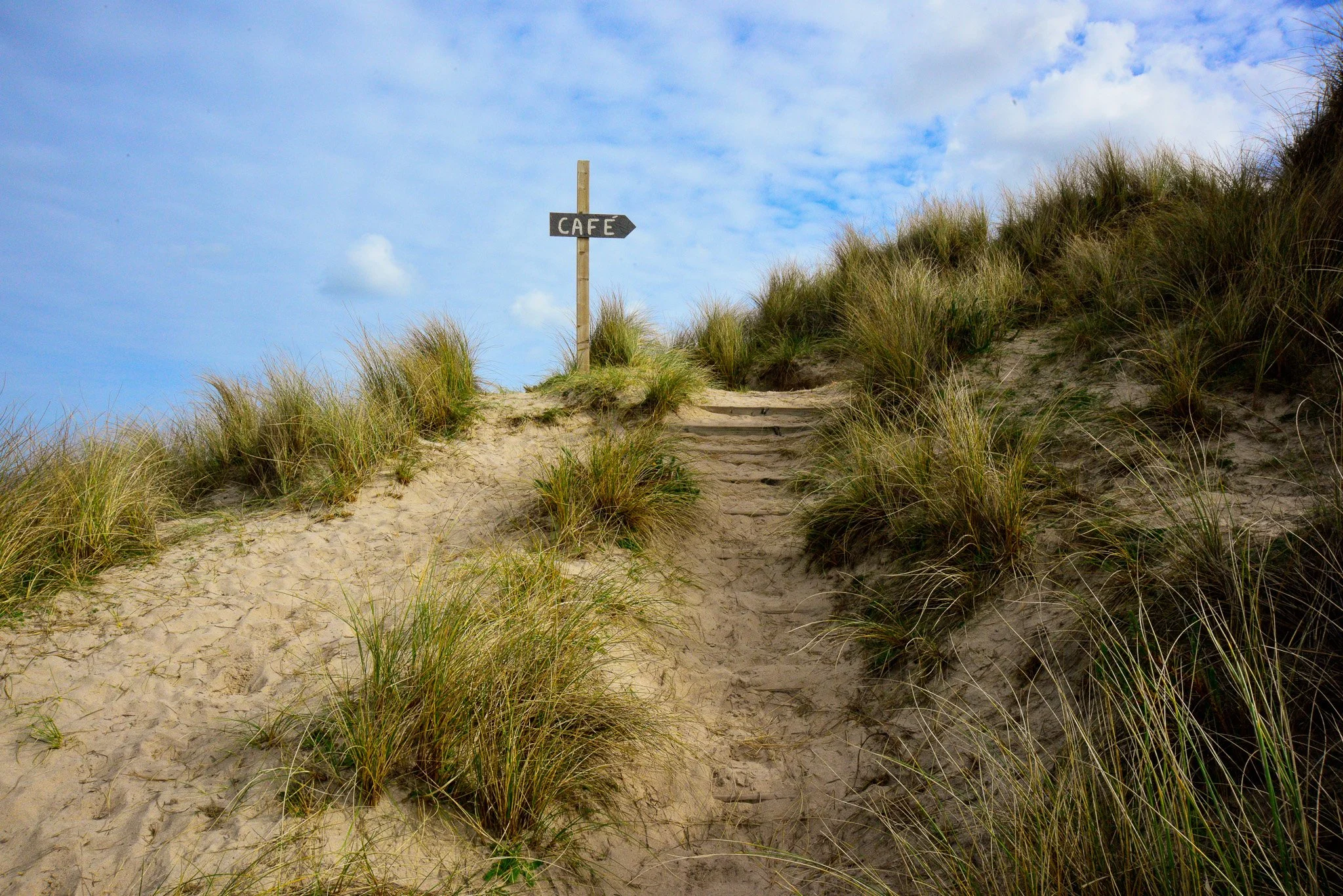 Sand dune with grassy plants and a wooden signpost indicating the direction to a cafe, under a partly cloudy sky.