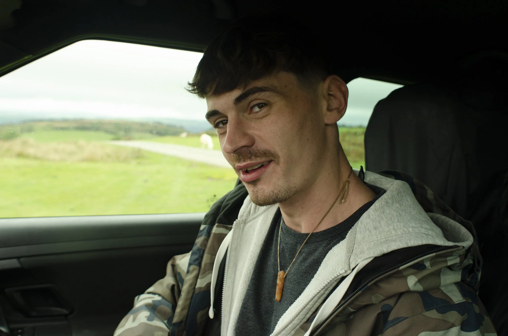 A young man with dark hair, a mustache, and a goatee sitting in the passenger seat of a vehicle, looking at the camera with a slight smile, wearing a camo jacket, a gray hoodie, and a necklace, with green fields and a cloudy sky visible through the w