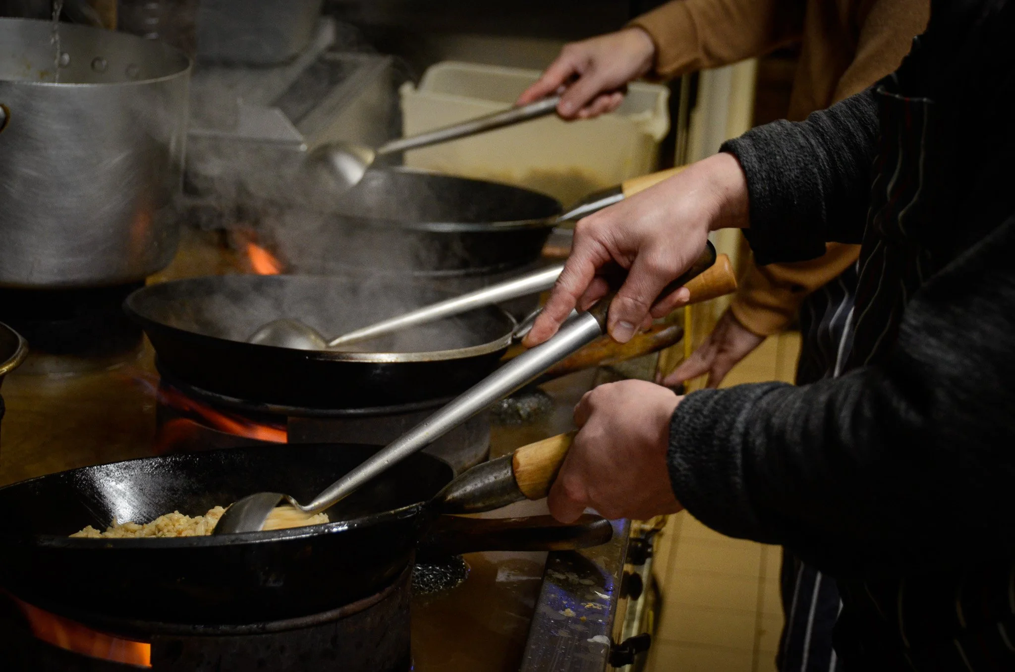 People cooking on a stovetop in a professional kitchen, stirring food in cast iron skillets over open flames.
