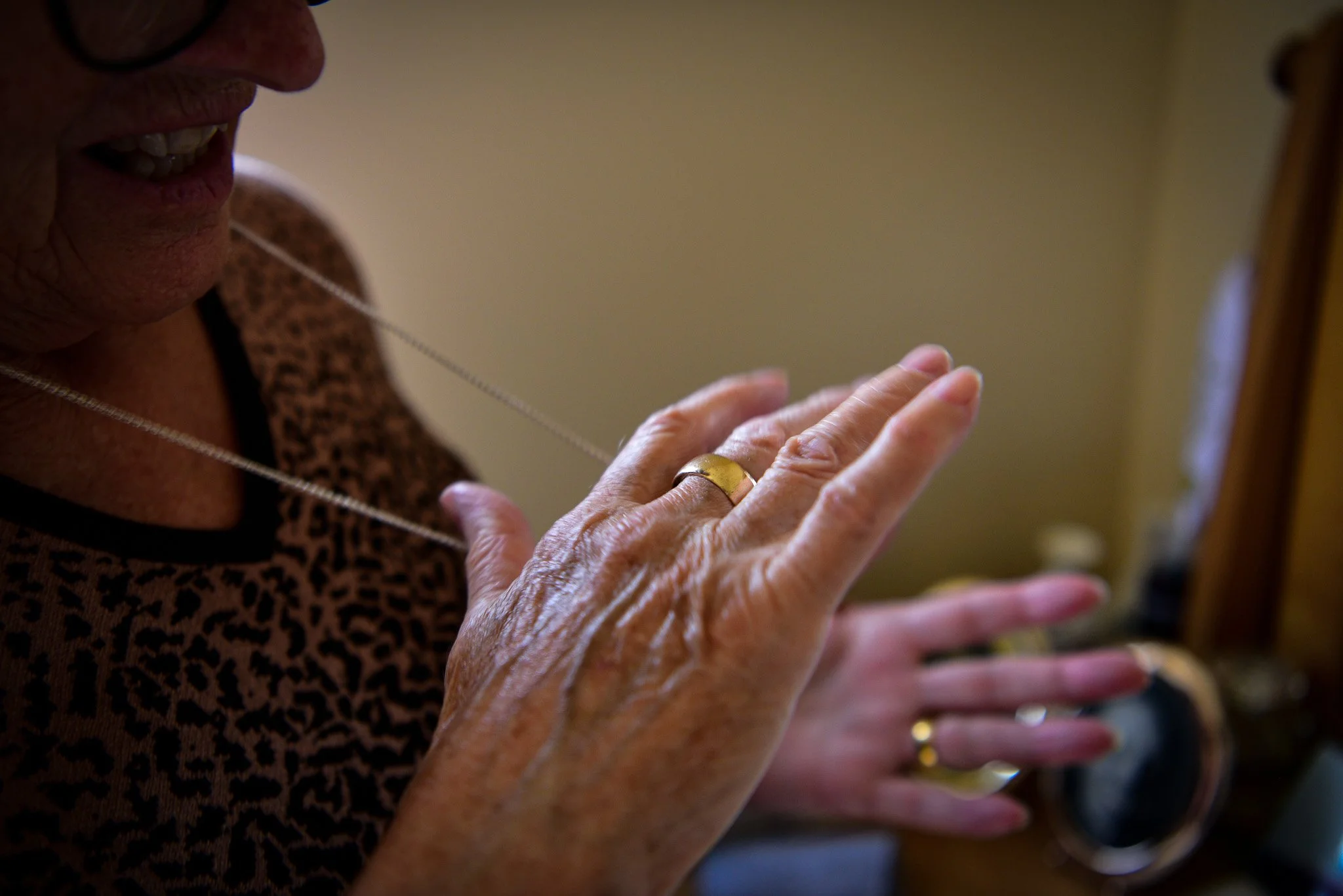 Close-up of an elderly woman wearing a leopard print top, looking at her hands with gold rings, with a mirror in the background.