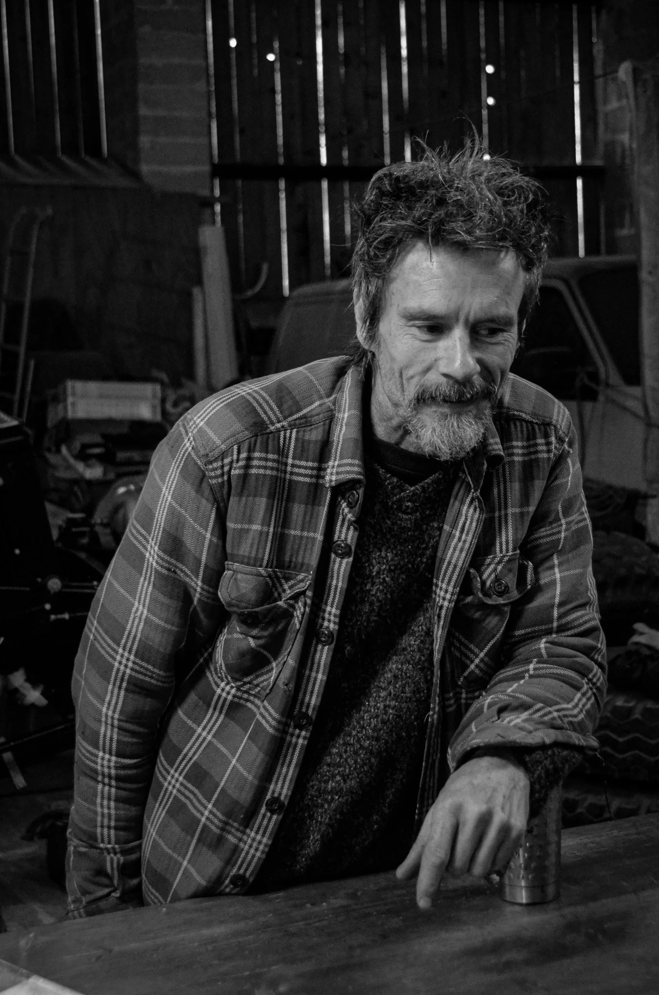 A man with curly hair and a beard wearing a plaid shirt and sweater, leaning on a wooden table in a workshop or garage, with tools and a vehicle in the background, in black and white.