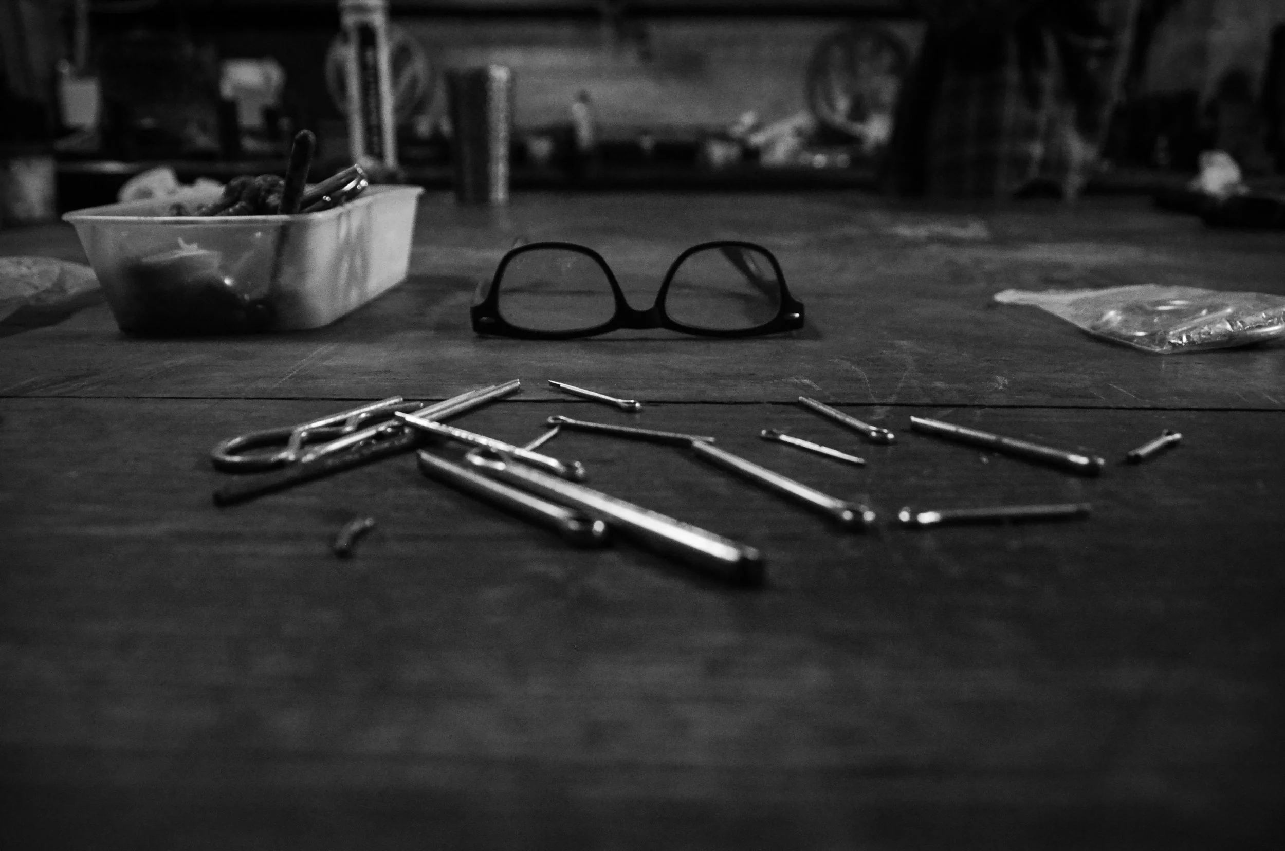 A black and white photo of a wooden table with scattered crochet hooks of various sizes, a pair of glasses, and a plastic container with some art supplies or snacks, in a workshop or craft space.