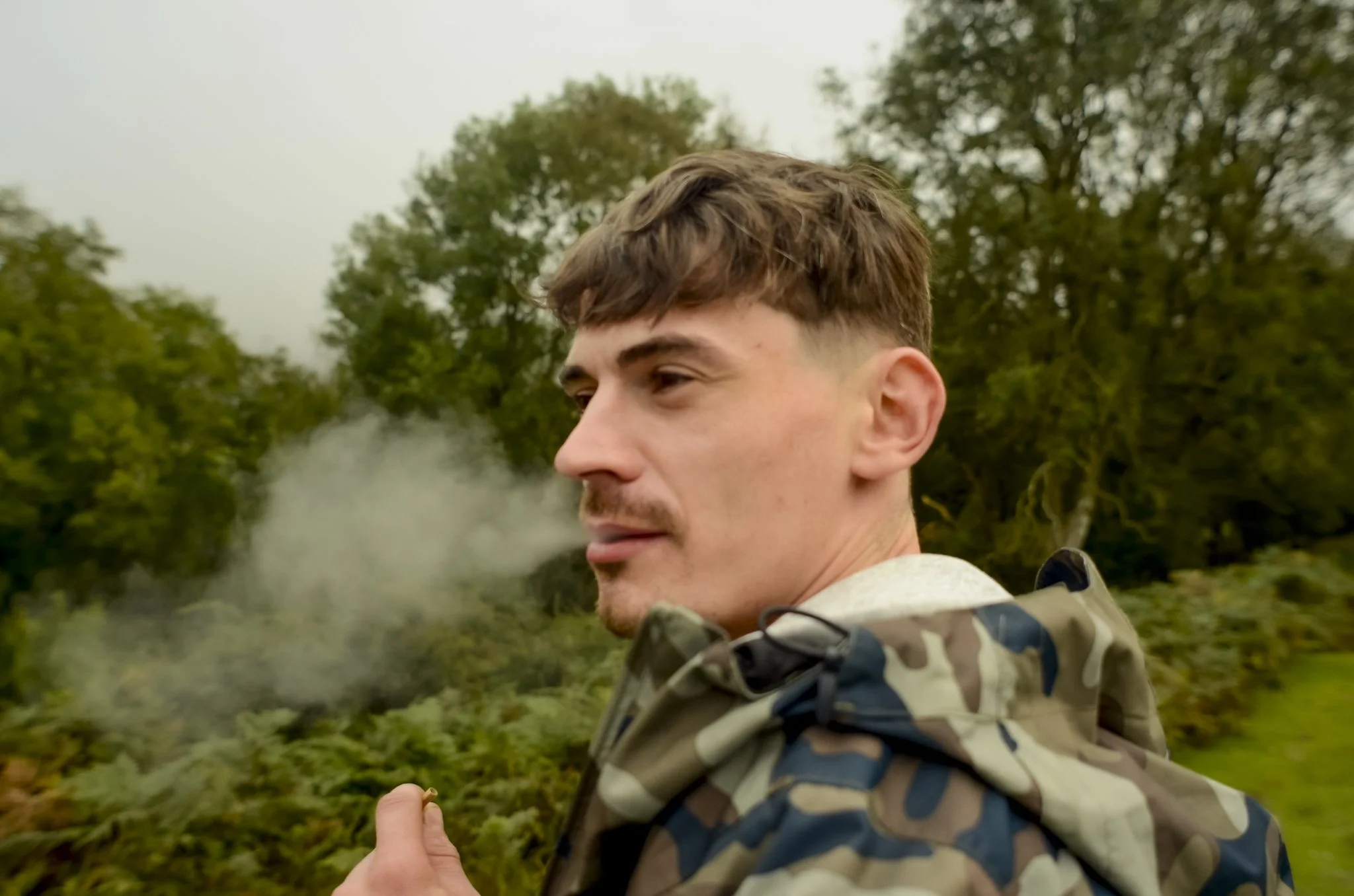 A young man with short brown hair and a mustache, wearing a camouflage jacket, standing outdoors in a natural setting with trees and bushes in the background, exhaling smoke.