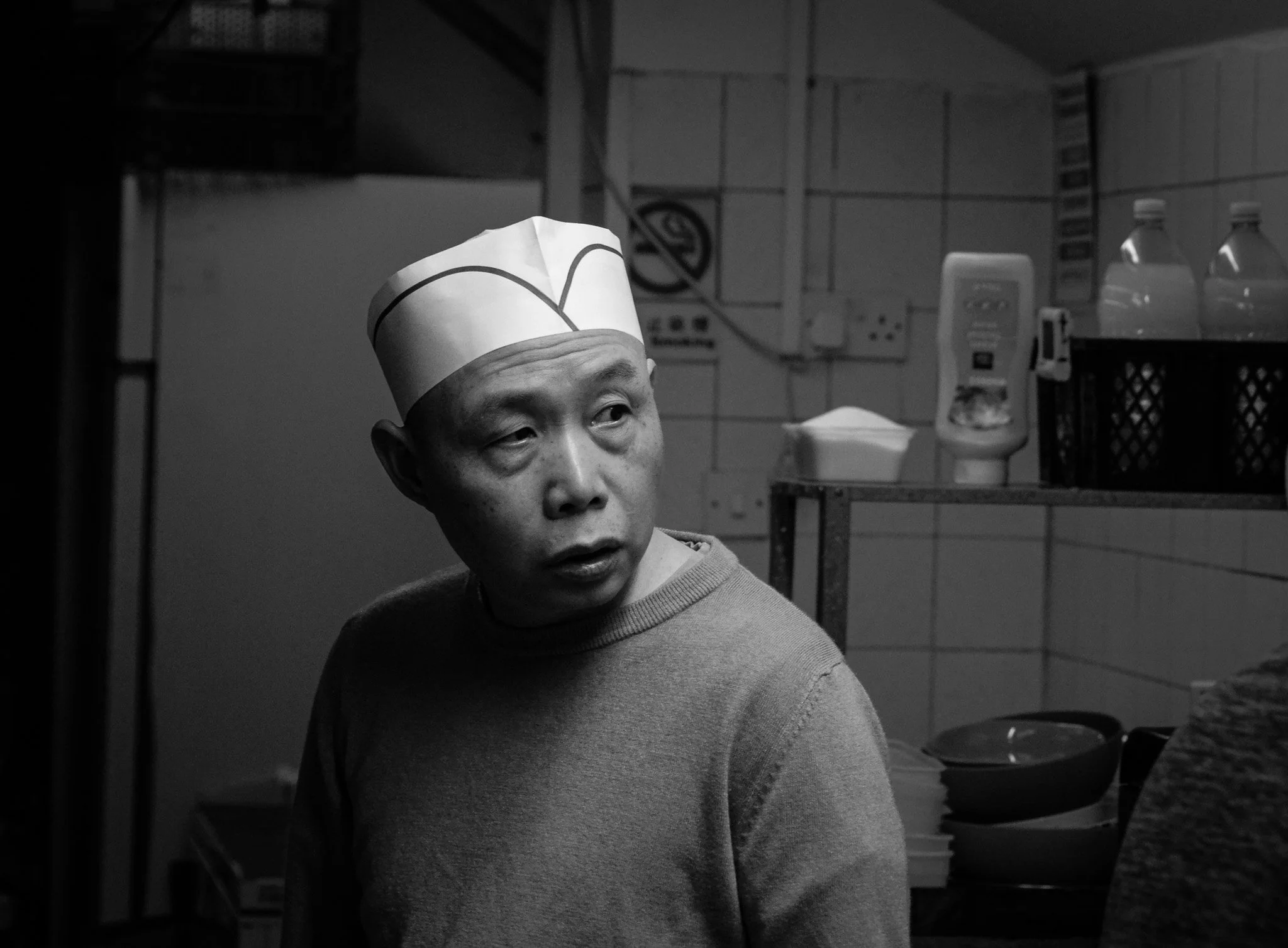 A black and white photo of a man wearing a paper chef's hat, looking to his left, in a kitchen with shelves, bottles, and containers.