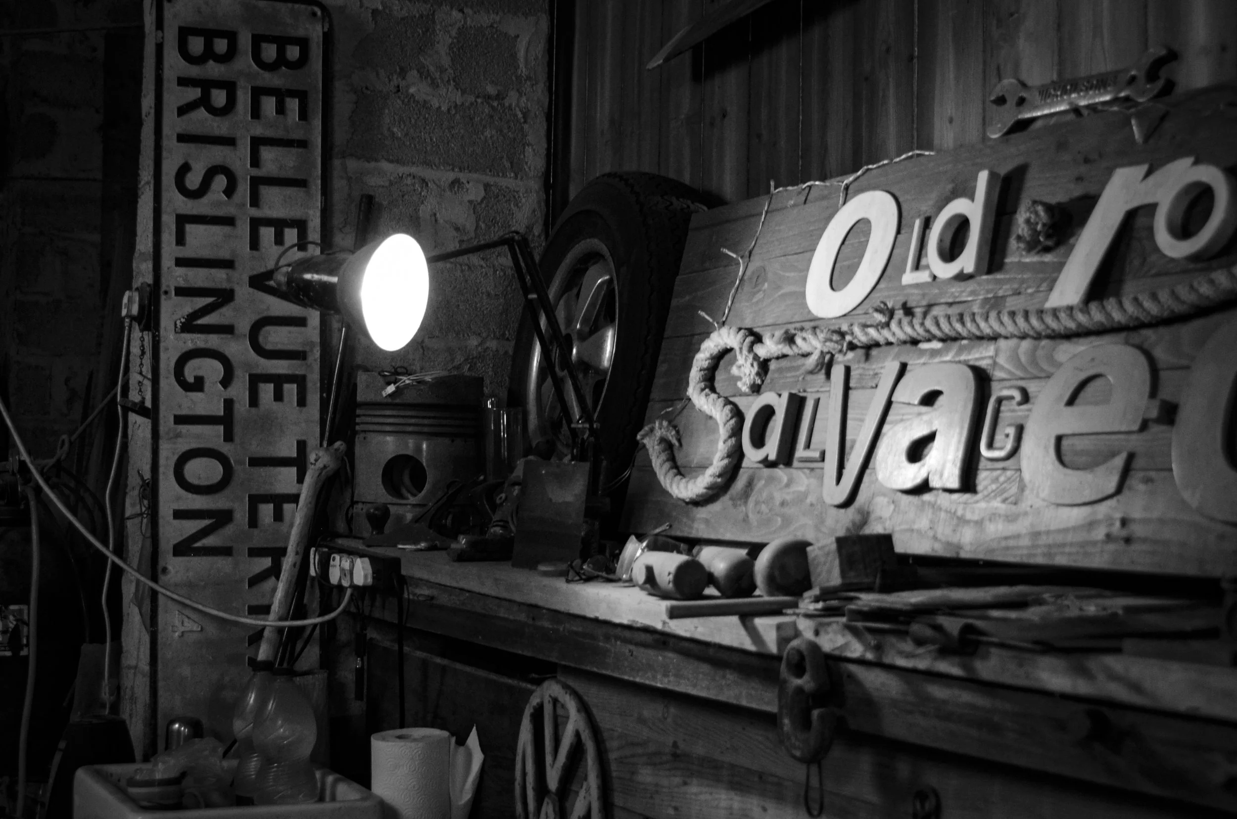 Black and white photo of a rustic workshop or garage with a wooden wall decorated with vintage signs and tools, including a large light, a bell, and miscellaneous items on shelves and the workbench.