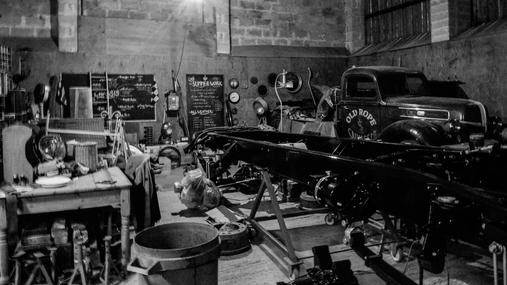 A cluttered garage or workshop with vintage items, including an old truck, a boat, tools, and furniture, with wall decorations and chalkboards.