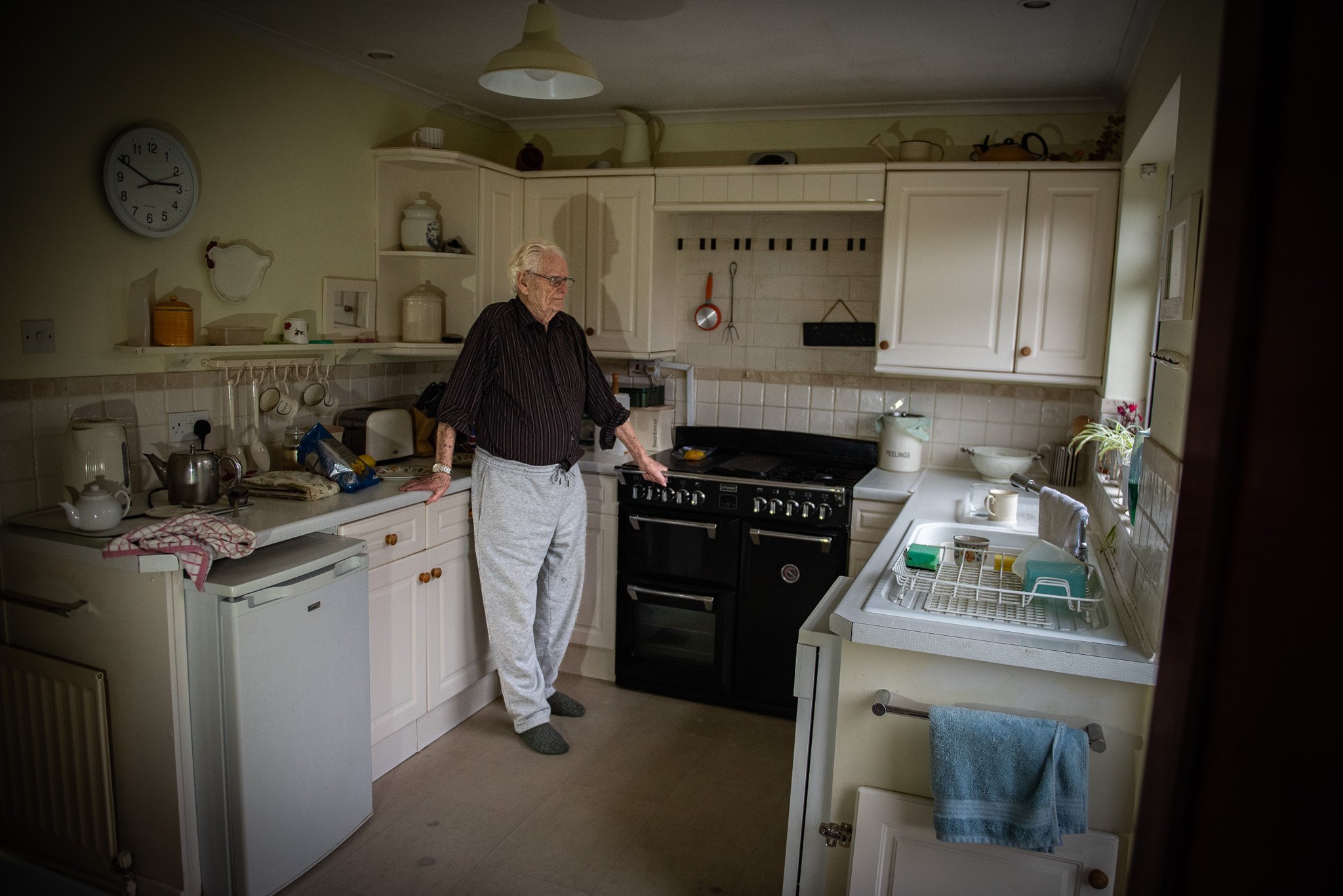 An elderly man with white hair and glasses stands in a kitchen, looking down at a black stove. The kitchen has white cabinets, a white tiled backsplash, and various kitchen items on the countertops. There is a clock on the wall and a towel hanging on