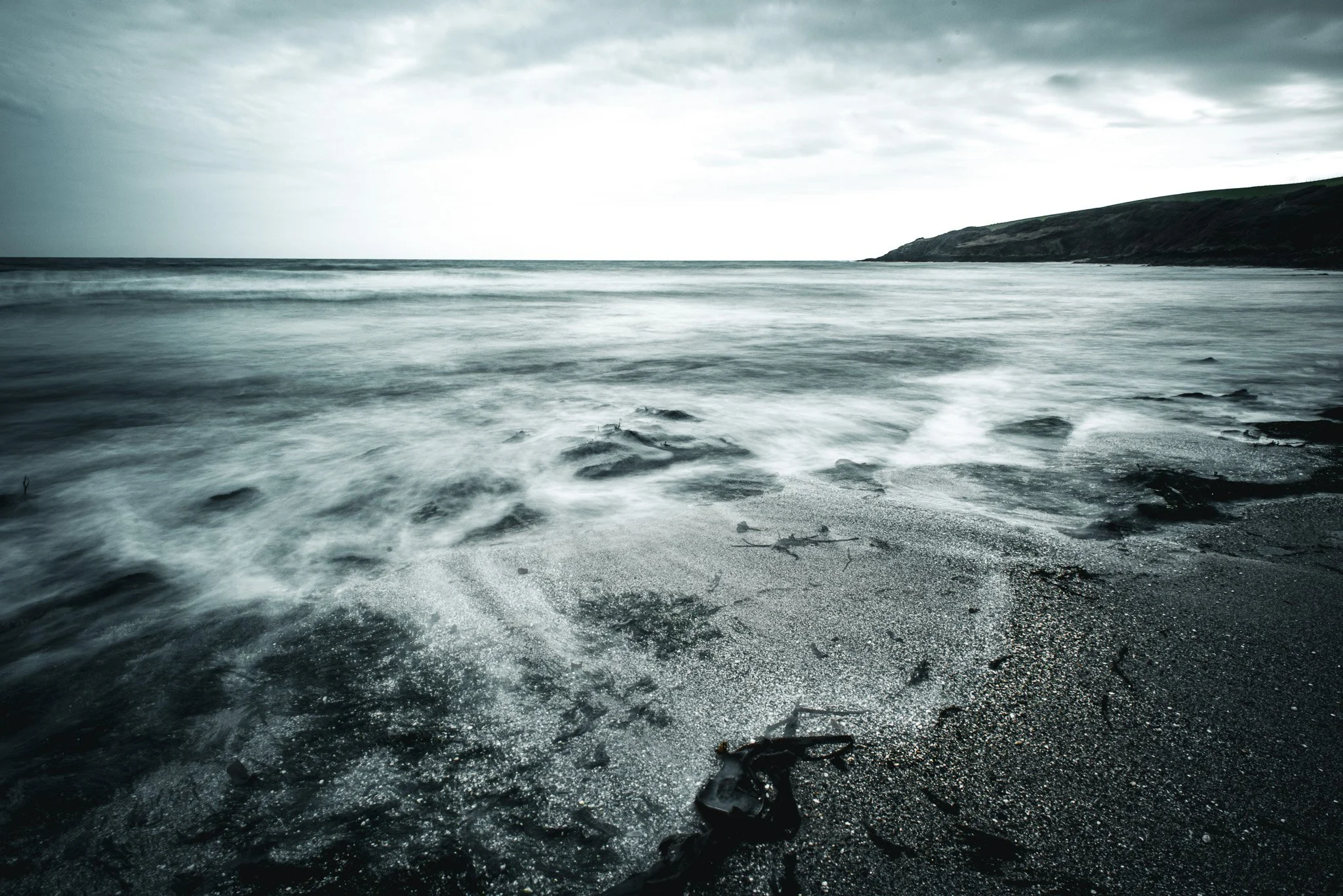 A black and white photograph of a beach with waves washing ashore, sand, rocks, and a distant shoreline under cloudy skies.