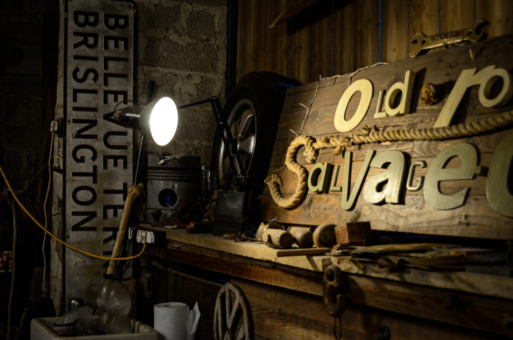 A vintage workshop with a wooden table holding tools, a large wooden sign that says 'Old lava,' a black car wheel, a lamp, and a metal sign reading 'Believe in your own direction' leaning against the wall.