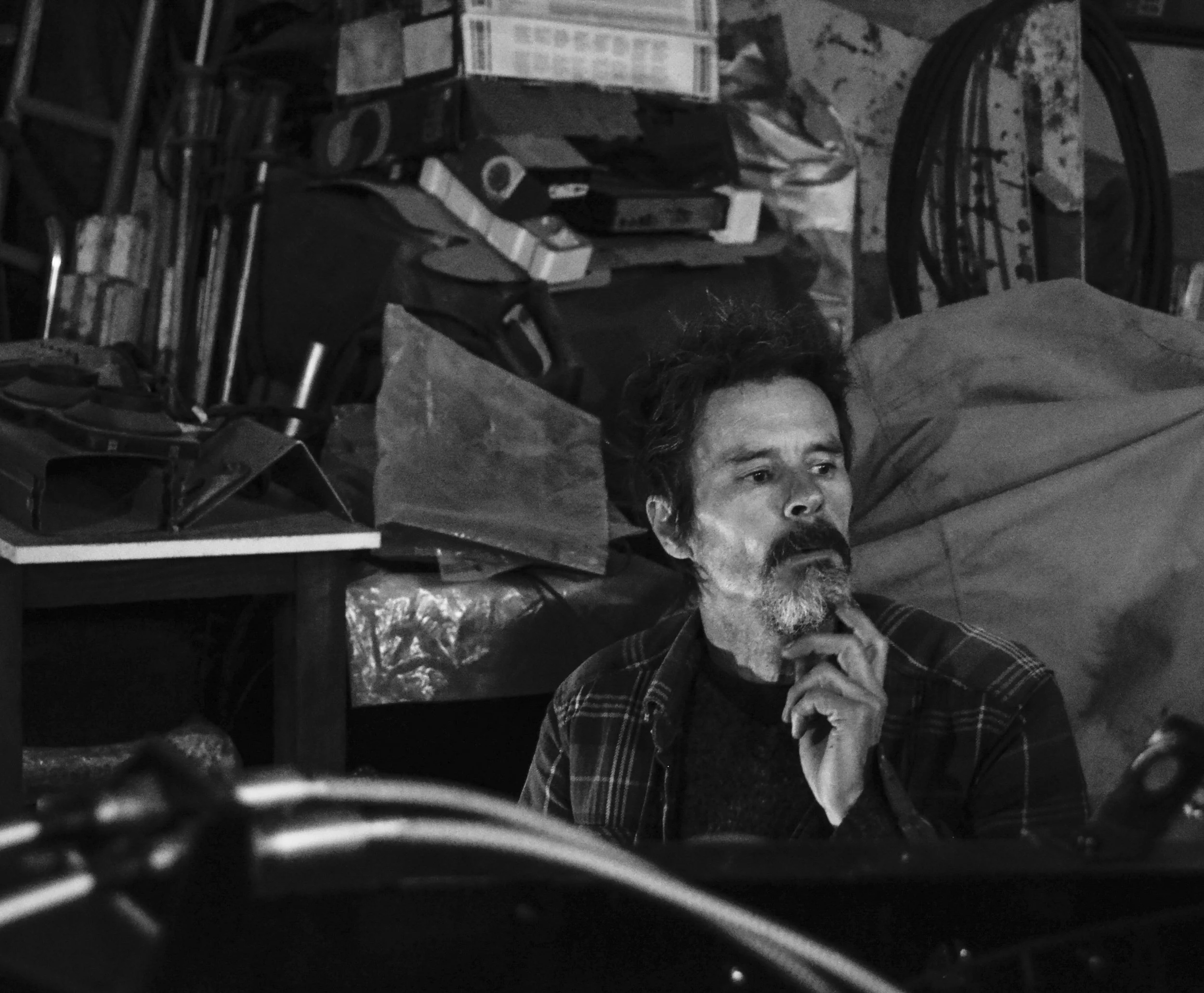 A man with a beard and mustache, wearing a plaid shirt, sitting in a cluttered room with various objects and boxes, appearing thoughtful with his hand on his chin.