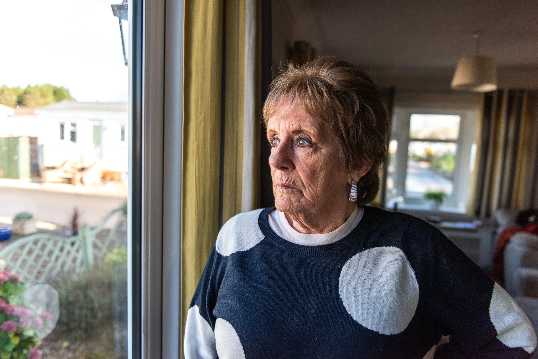 An older woman with short light brown hair looking out a window, with indoor lighting and a blurred background of a room and outside view.