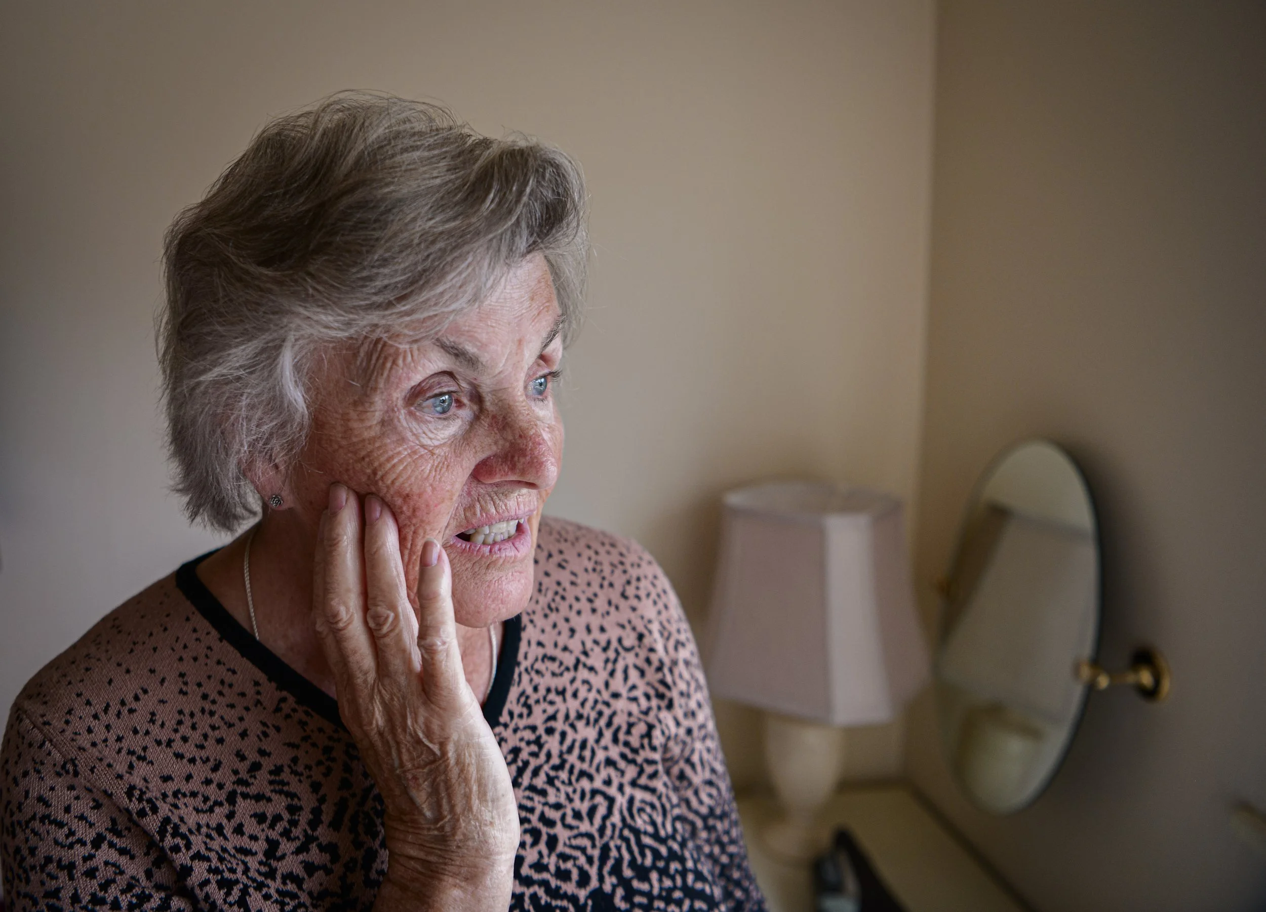 An elderly woman with gray hair and blue eyes touching her face, looking concerned or worried, with a mirror and a lamp in the background.