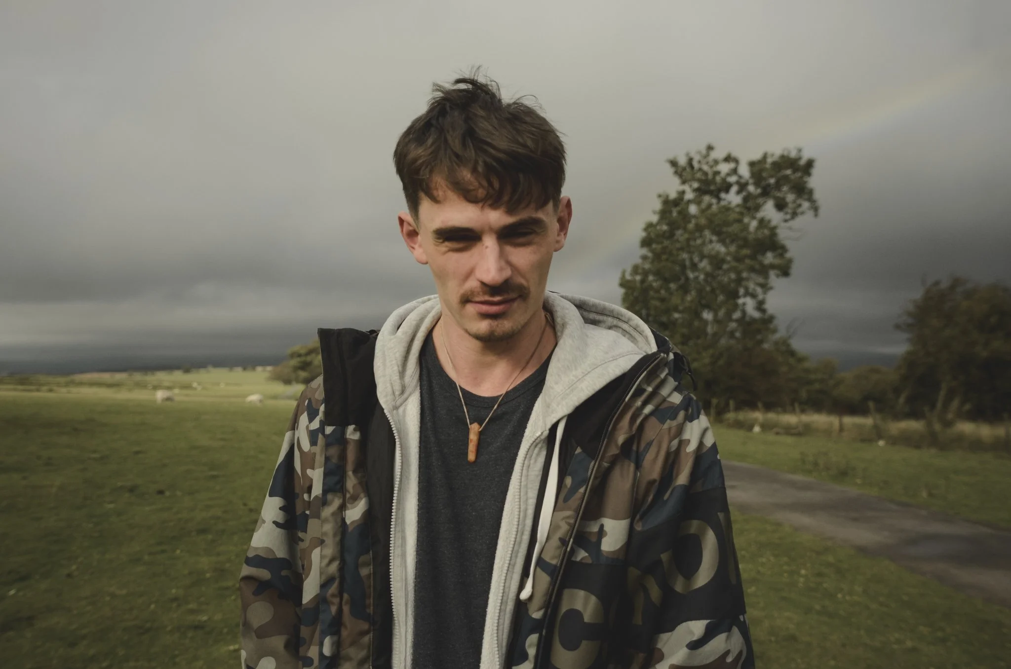 Young man standing outdoors on a cloudy day with a rainbow in the background, wearing a camouflage jacket, gray hoodie, and a necklace.